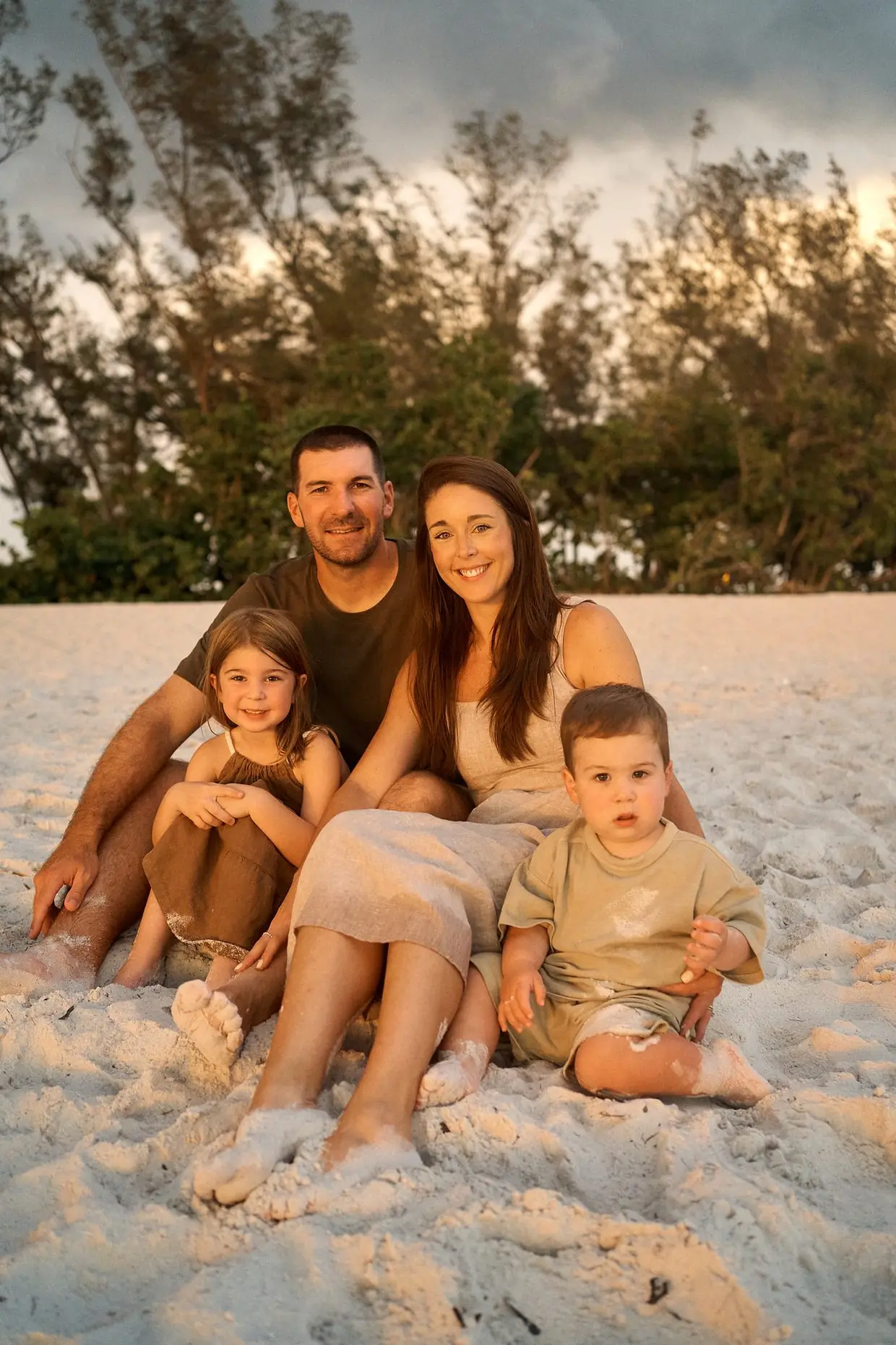 A mother, a father, a little girl in a brown dress and a toddler sitting close together on white sand on Anna Maria Island, warm low sun on their faces and a heavy dark grey sky behind the trees