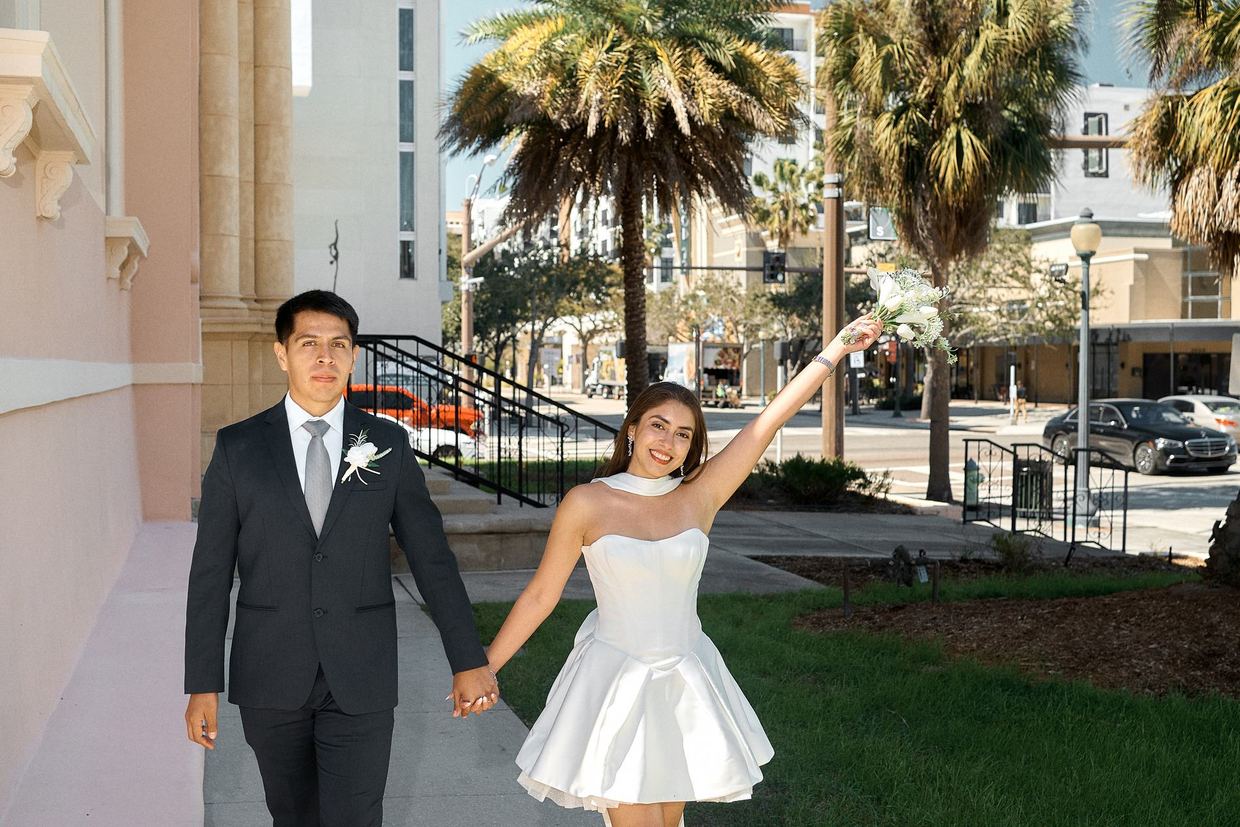 A bride raising her arm in joy as she walks with her new husband past the Sarasota Historic Courthouse