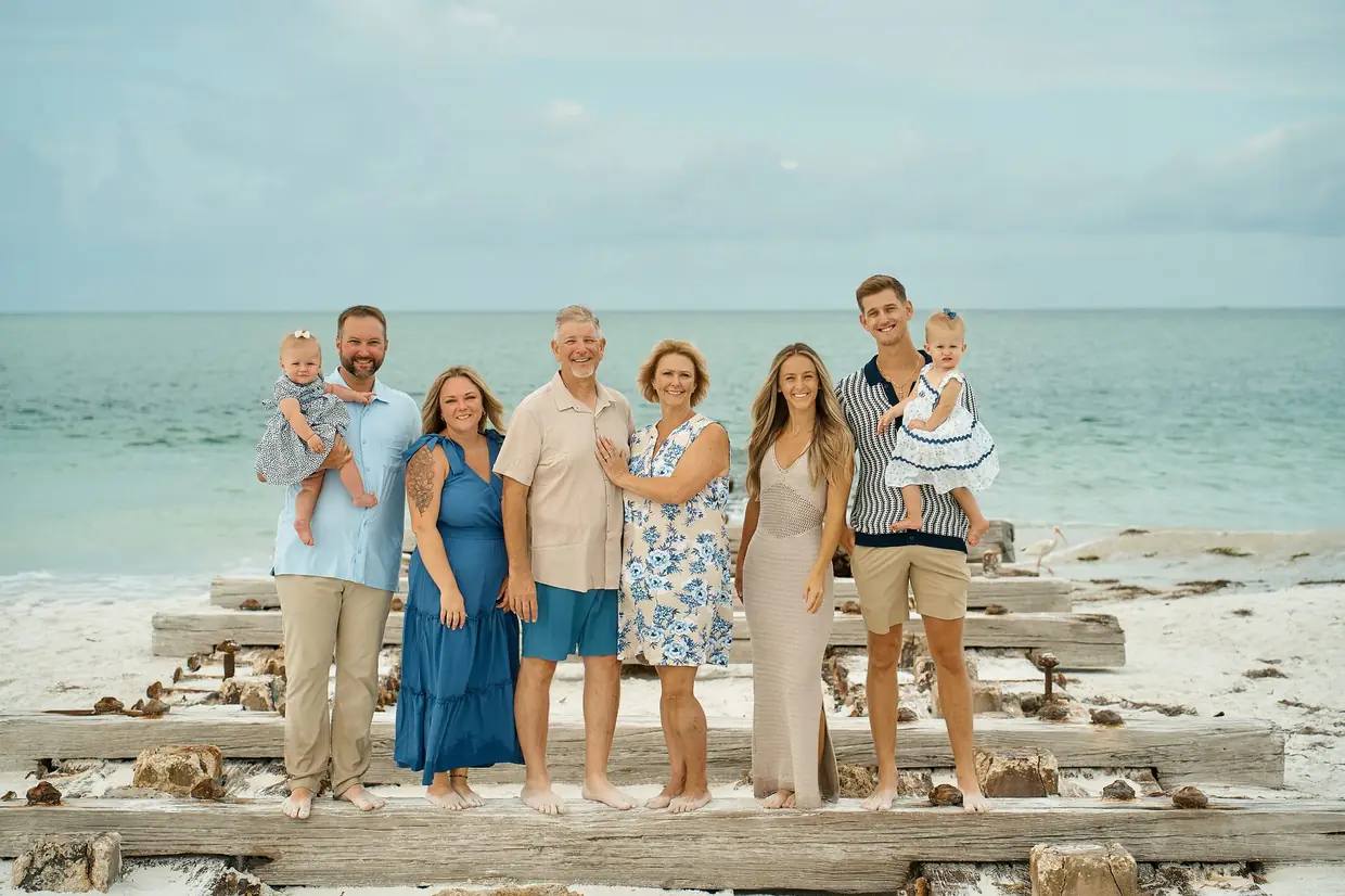 A family of eight, three generations with two baby girls, standing in a line on weathered timbers on the sand, the green Gulf behind them