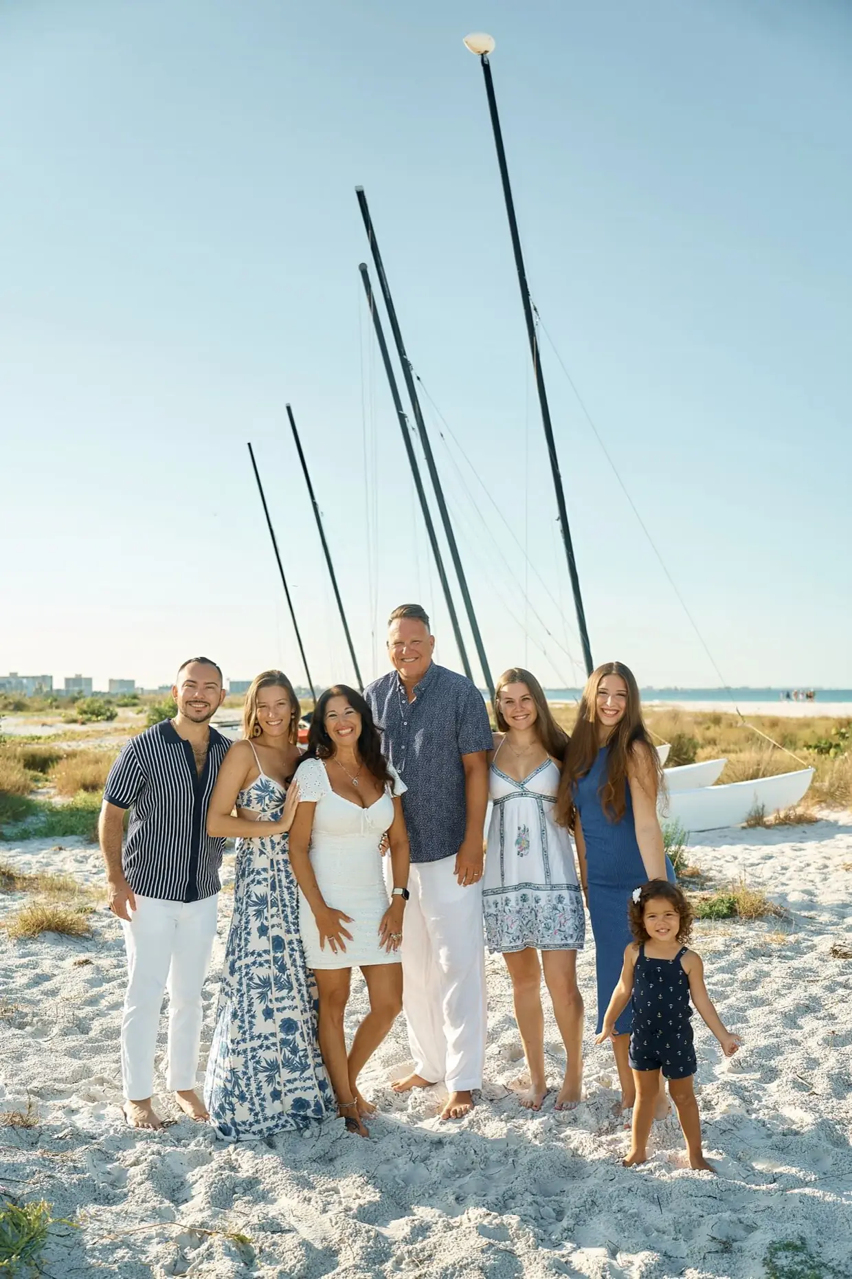 A family of seven in blue and white standing close together on the white sand of Siesta Key, a little girl in a navy romper in front, catamaran masts behind them