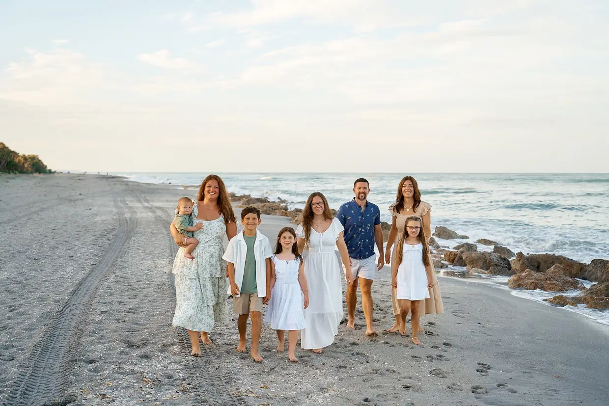 A family group of eight in white, sage, beige and blue, from a baby in arms to the grown-ups, walking together along the water at Caspersen Beach in Venice with rocks in the surf beside them