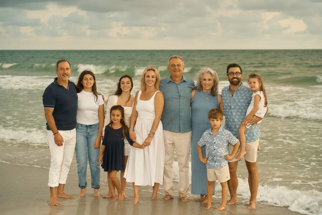 A family of ten, grandparents, their grown children and grandchildren from a toddler to teenagers, standing together barefoot at the edge of the sea under a soft cloudy sky