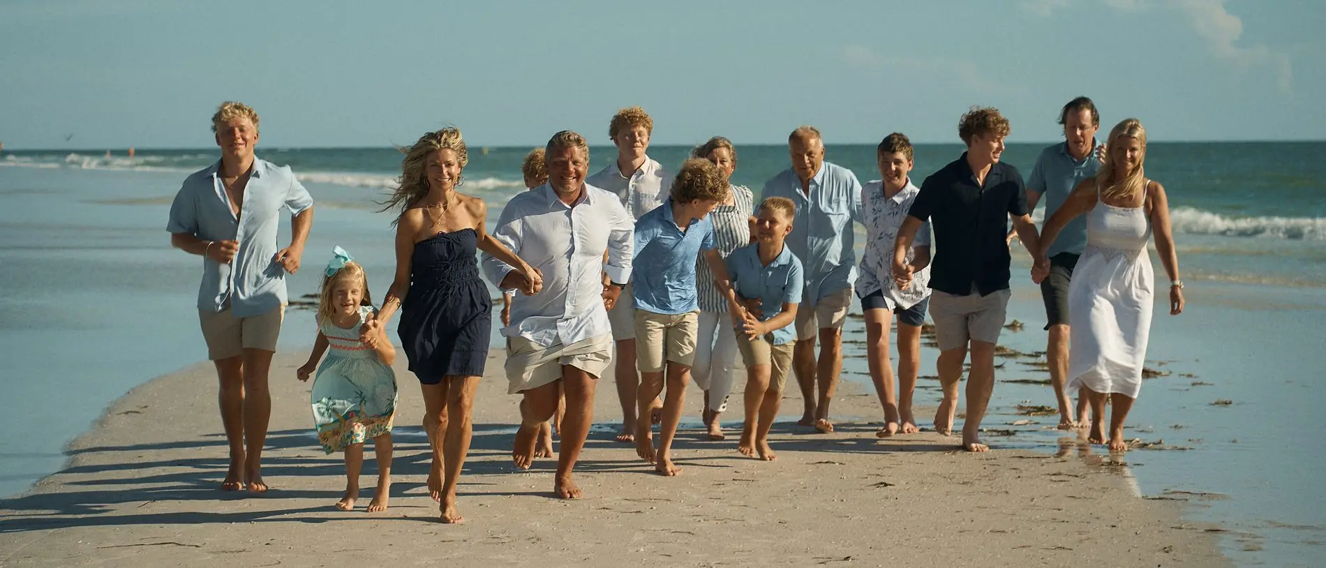 A big family of about fourteen, three generations, running barefoot toward the camera along a wet sandbar, the children out in front and the Gulf behind them