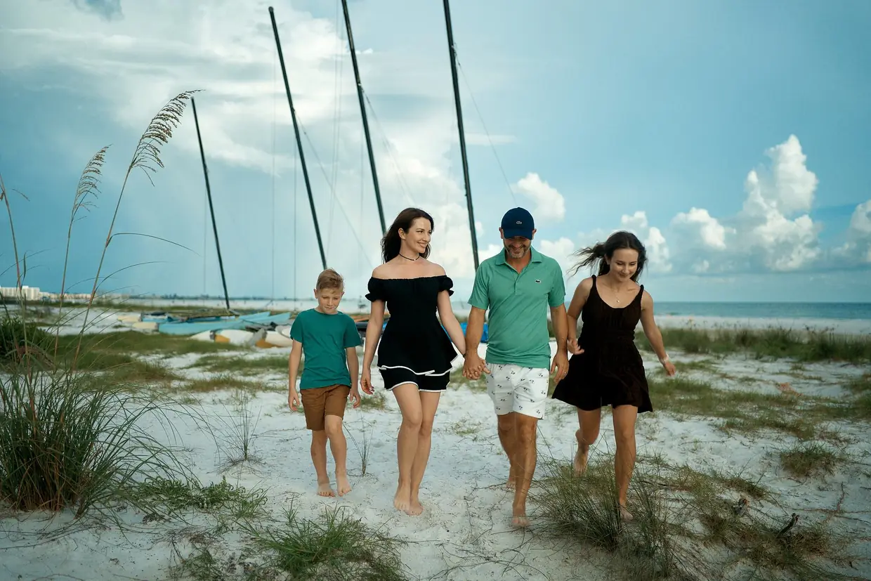 A family of four walking toward the camera through dune grass on white sand, a teenage girl and a young boy with their parents, catamaran masts and big white clouds behind them