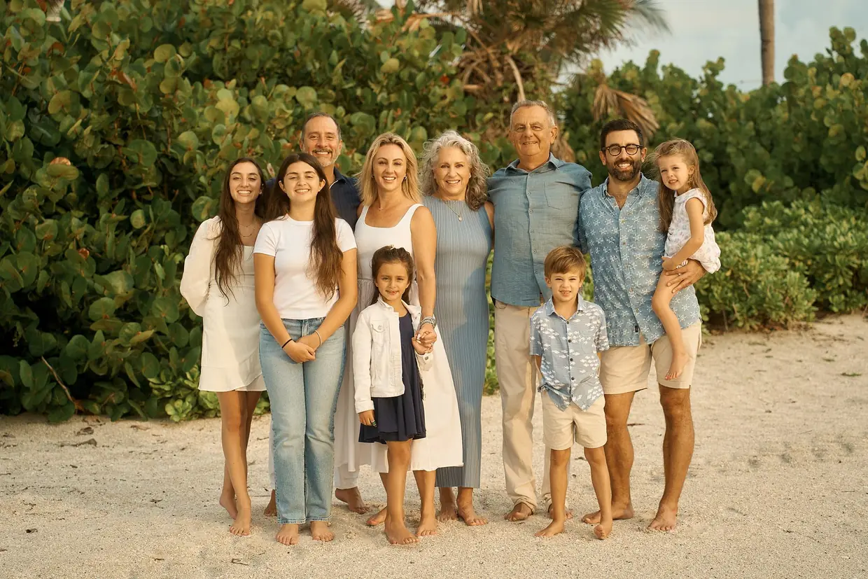 Three generations of one family, ten people, standing together on white sand in front of a wall of sea grape: the grandparents in the middle, their grown children around them, a little girl on her father's hip and a boy in front