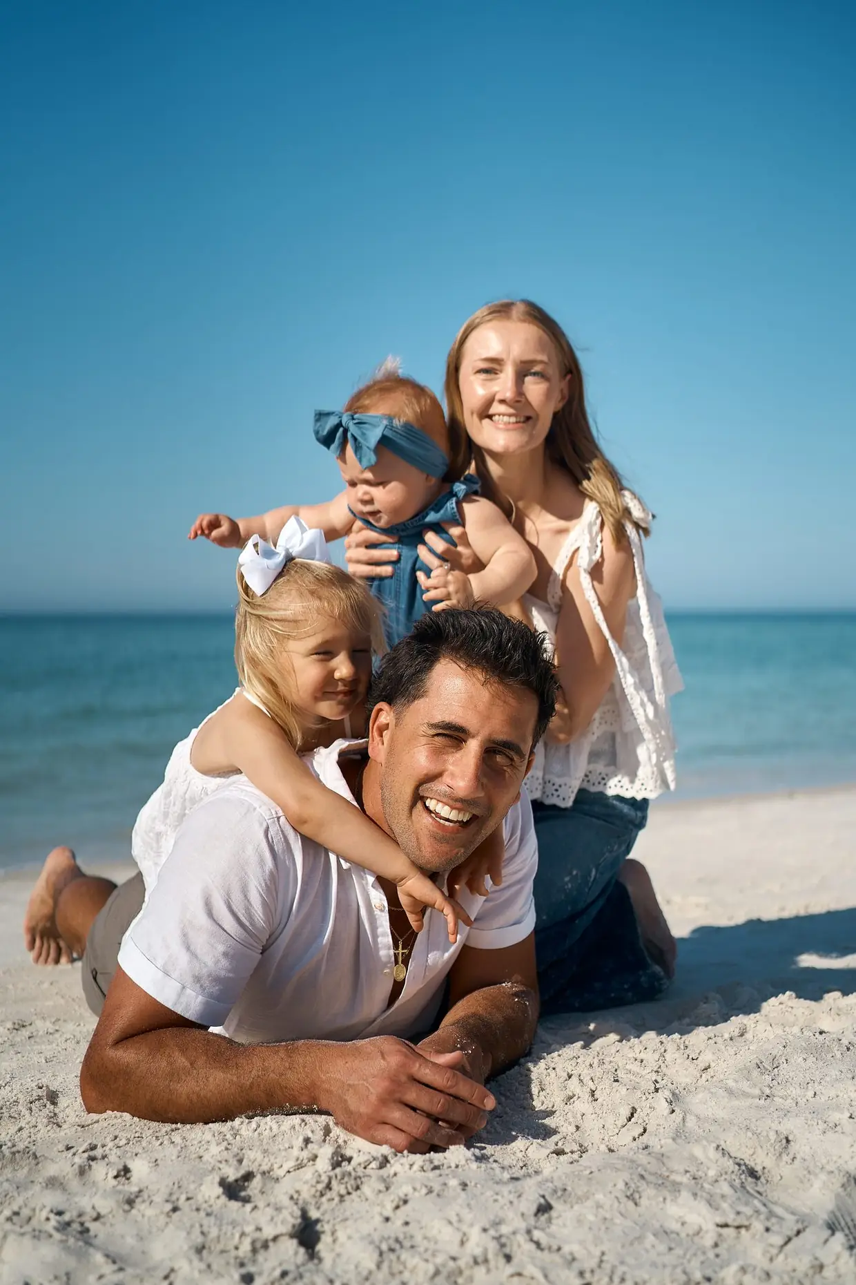 A laughing father lying on the sand with his little girl on his back, the mother behind him holding a baby in a blue bow, on Siesta Key under a clear blue sky