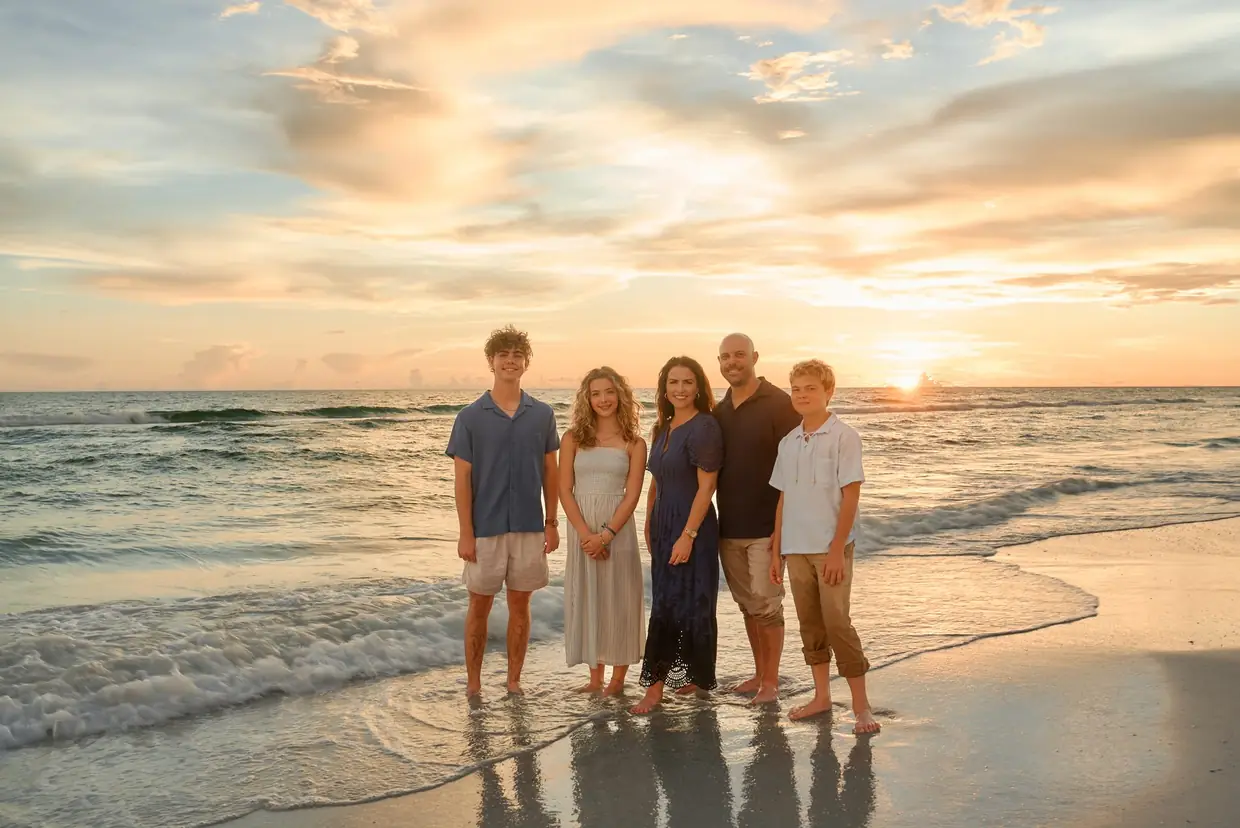 A family of five standing at the edge of the water on Siesta Key, the sun touching the Gulf horizon behind them