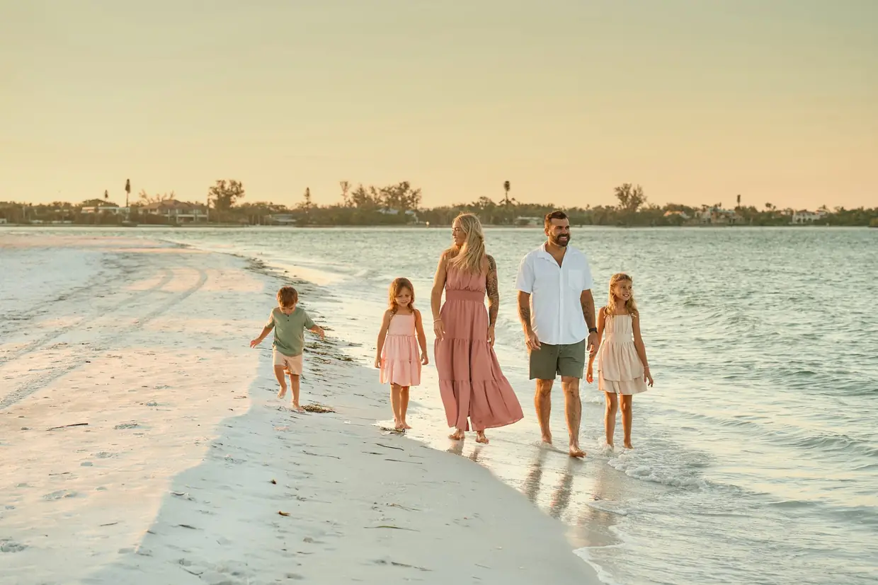 A family of five walking along a white sandbar at the edge of the water on South Lido Beach in Sarasota, the mother in a long pink dress, the father in a white shirt, two little girls in pastel dresses and their little brother walking ahead in warm evening light