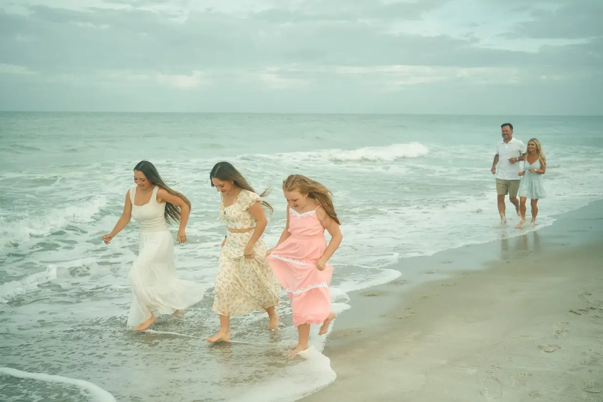 Three sisters in white, floral and pink dresses running through the surf on the beach at The St. Regis Longboat Key, their parents walking hand in hand behind them