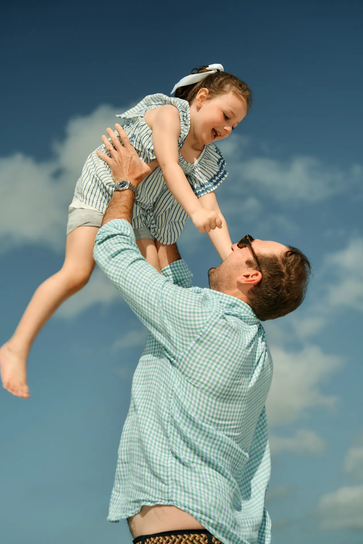 A father lifting his laughing daughter high above his head against a blue sky with white clouds on Anna Maria Island