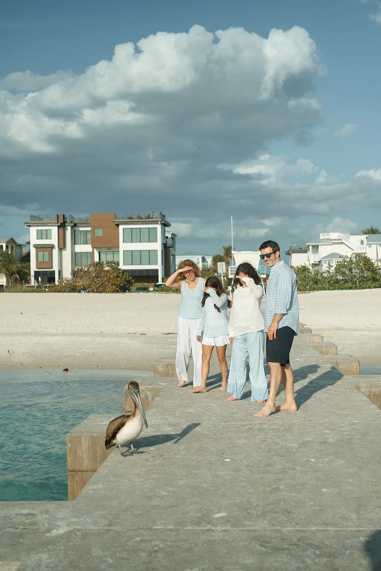 A family of five on a concrete jetty on Anna Maria Island, one woman holding her hair in the wind, a pelican in the foreground and huge white clouds over the houses