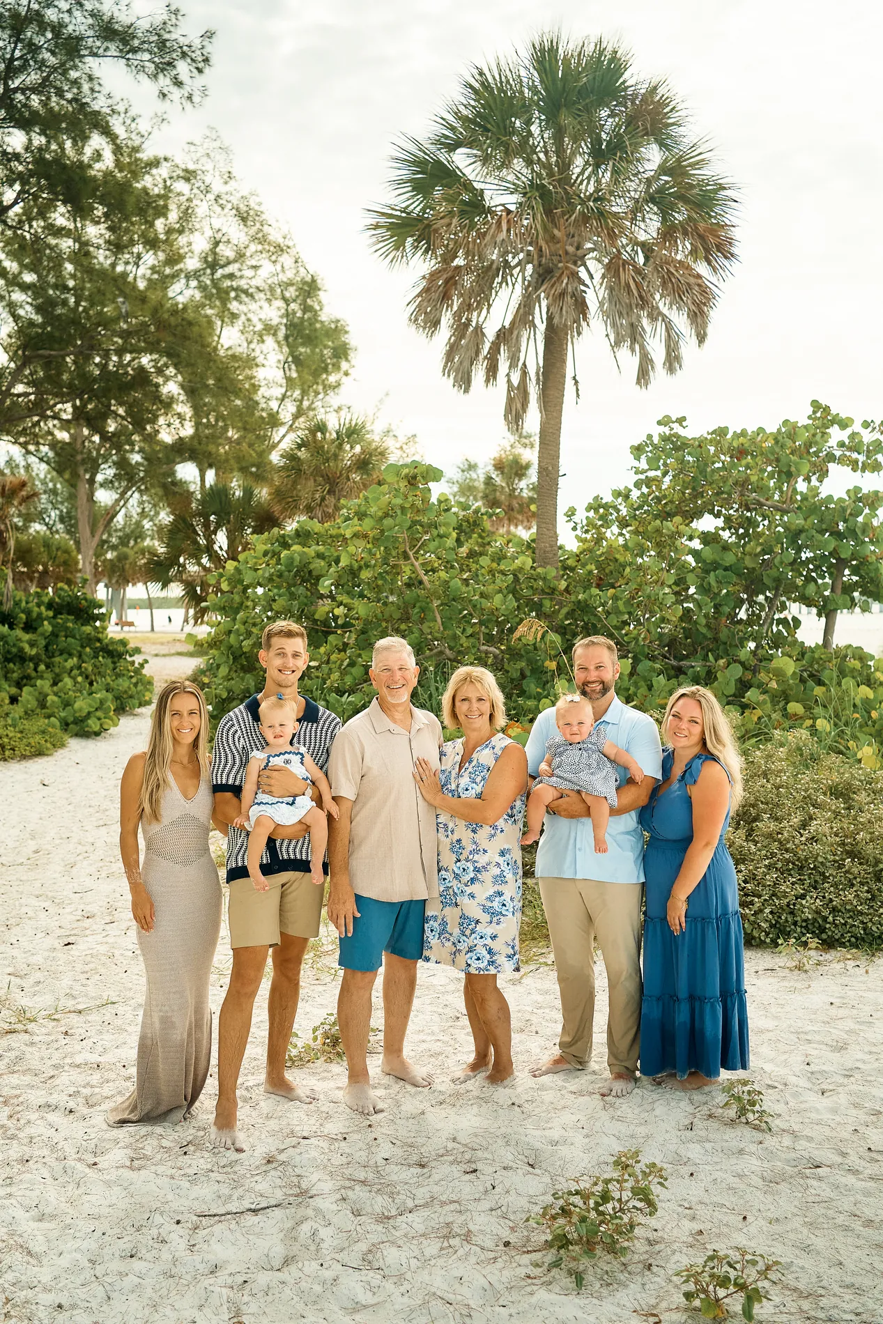 Three generations, eight people, on Anna Maria Island: the grandparents in the middle, two fathers each holding a baby, the women in long blue and beige dresses, a tall palm and green trees behind them