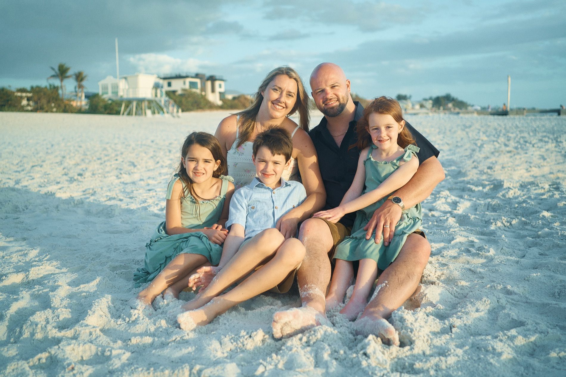 A family of five sitting together on the sand on Anna Maria Island, beach cottages behind them