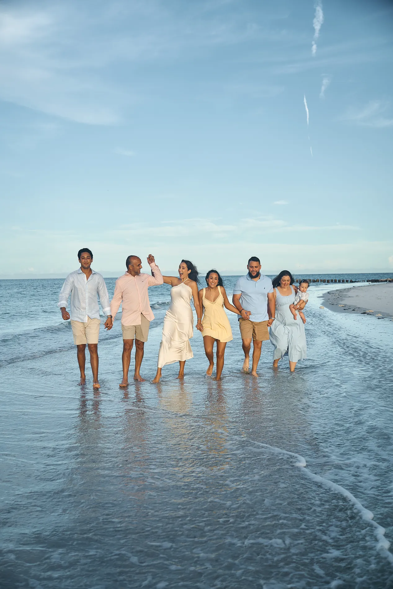 Six adults walking hand in hand toward the camera through the shallows on Anna Maria Island, a mother on the right carrying a baby, under a clear blue sky