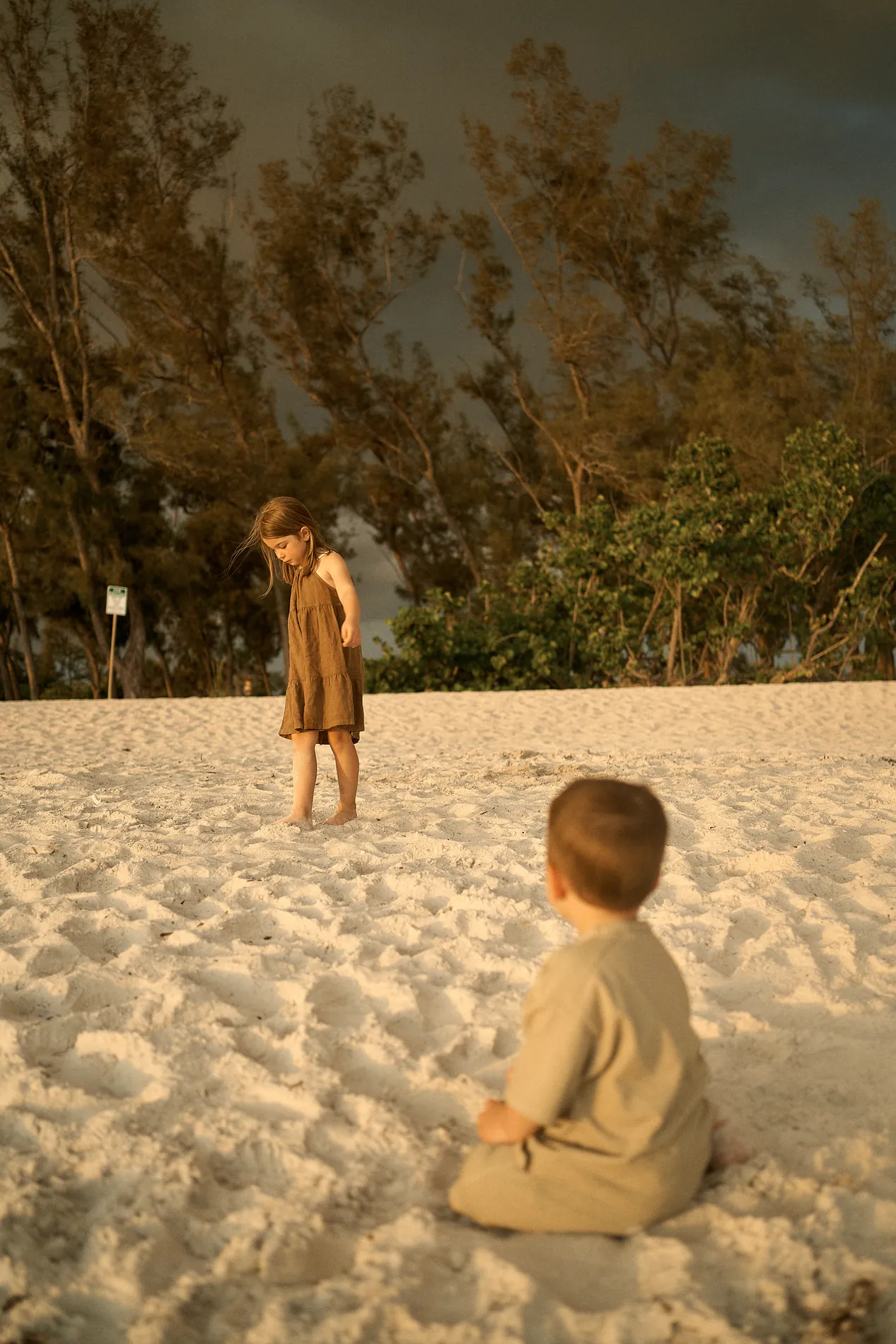 A little girl in a brown dress standing barefoot on white sand on Anna Maria Island and her small brother sitting in the sand watching her, warm low light on them and an almost black sky behind the trees
