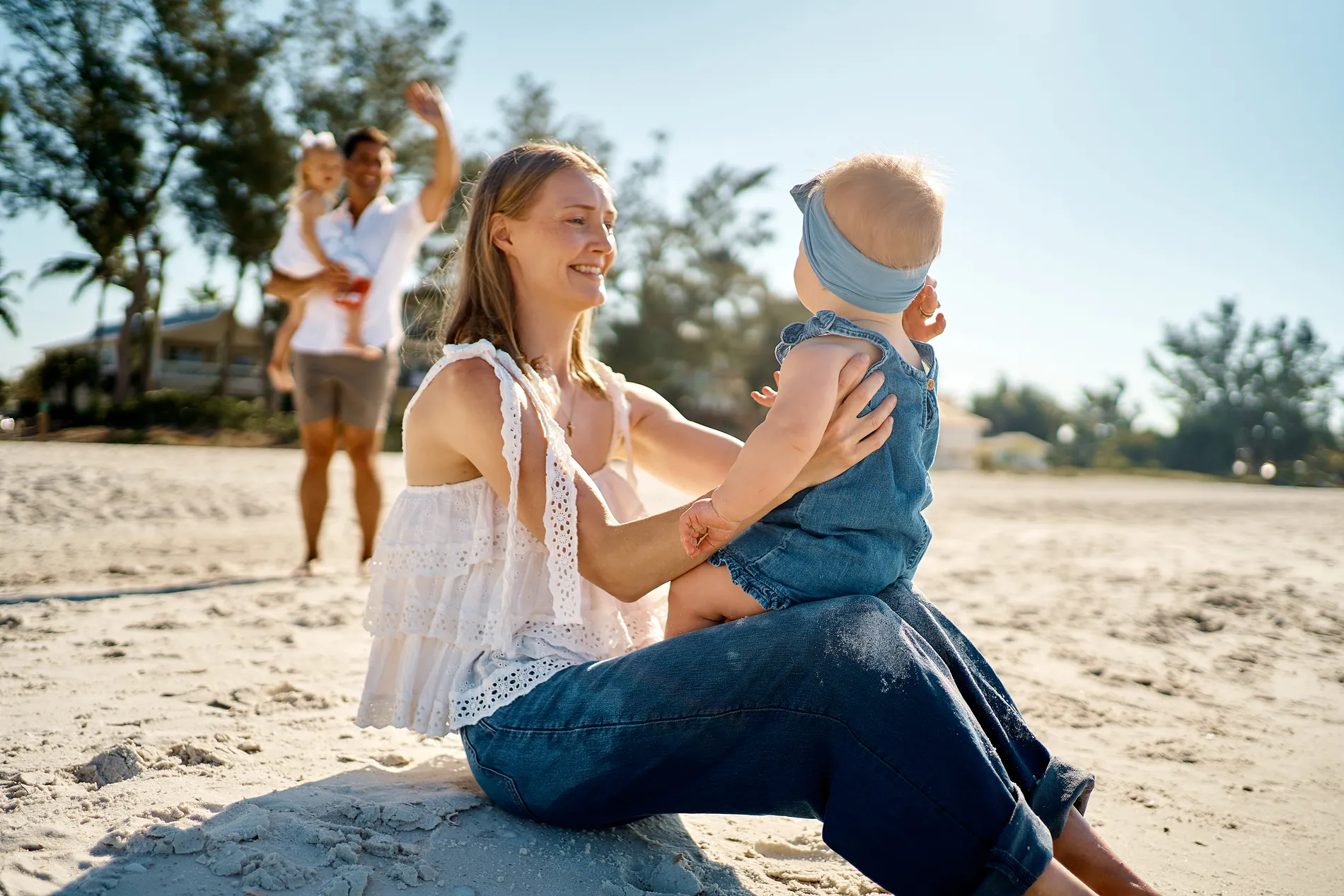 A mother in a white lace top and jeans sitting on the sand on Anna Maria Island, laughing with her baby in a denim romper and blue headband, the father and an older child behind them