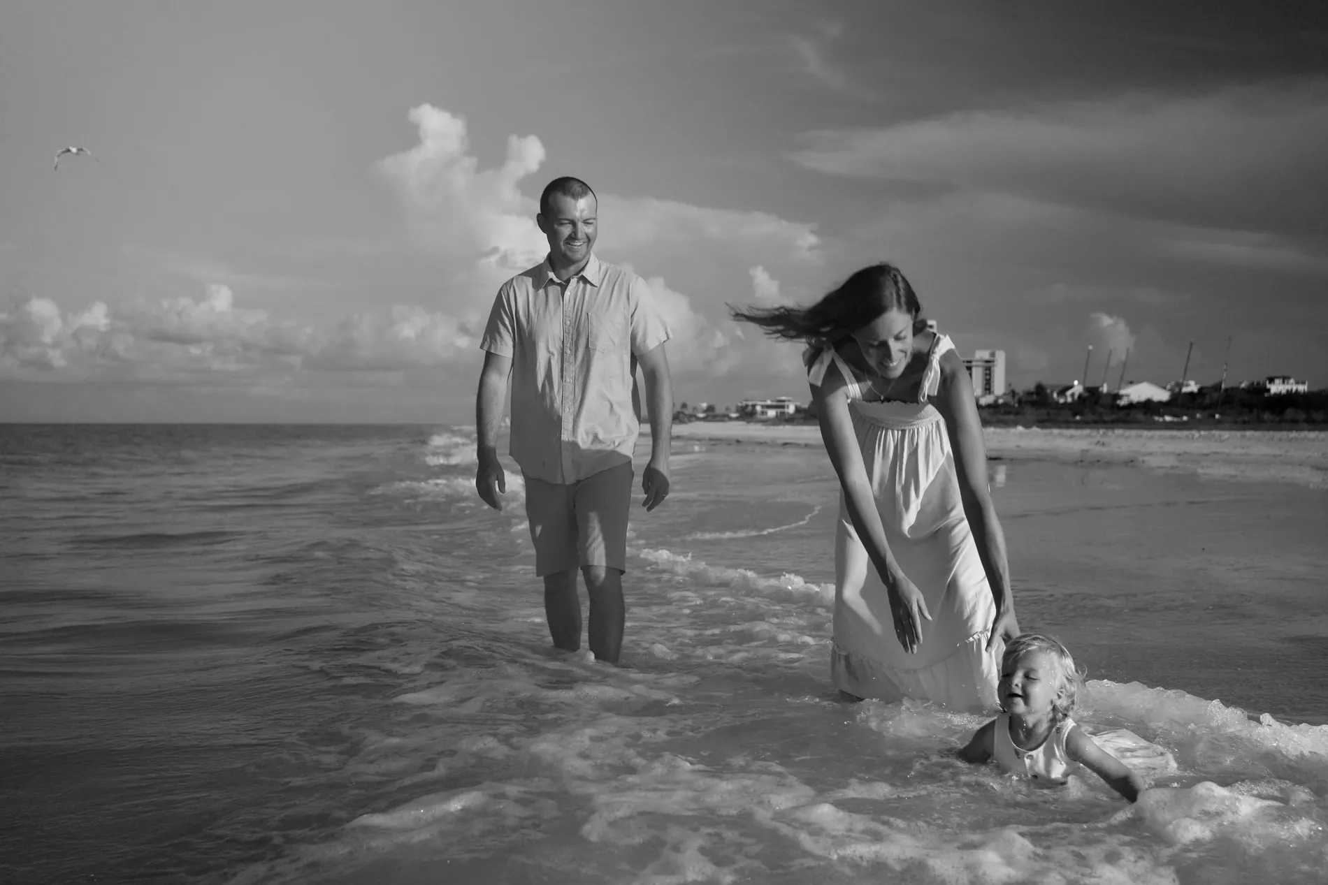 Black and white: a father walking through the shallow water while his daughter bends down to a small child splashing at the edge
