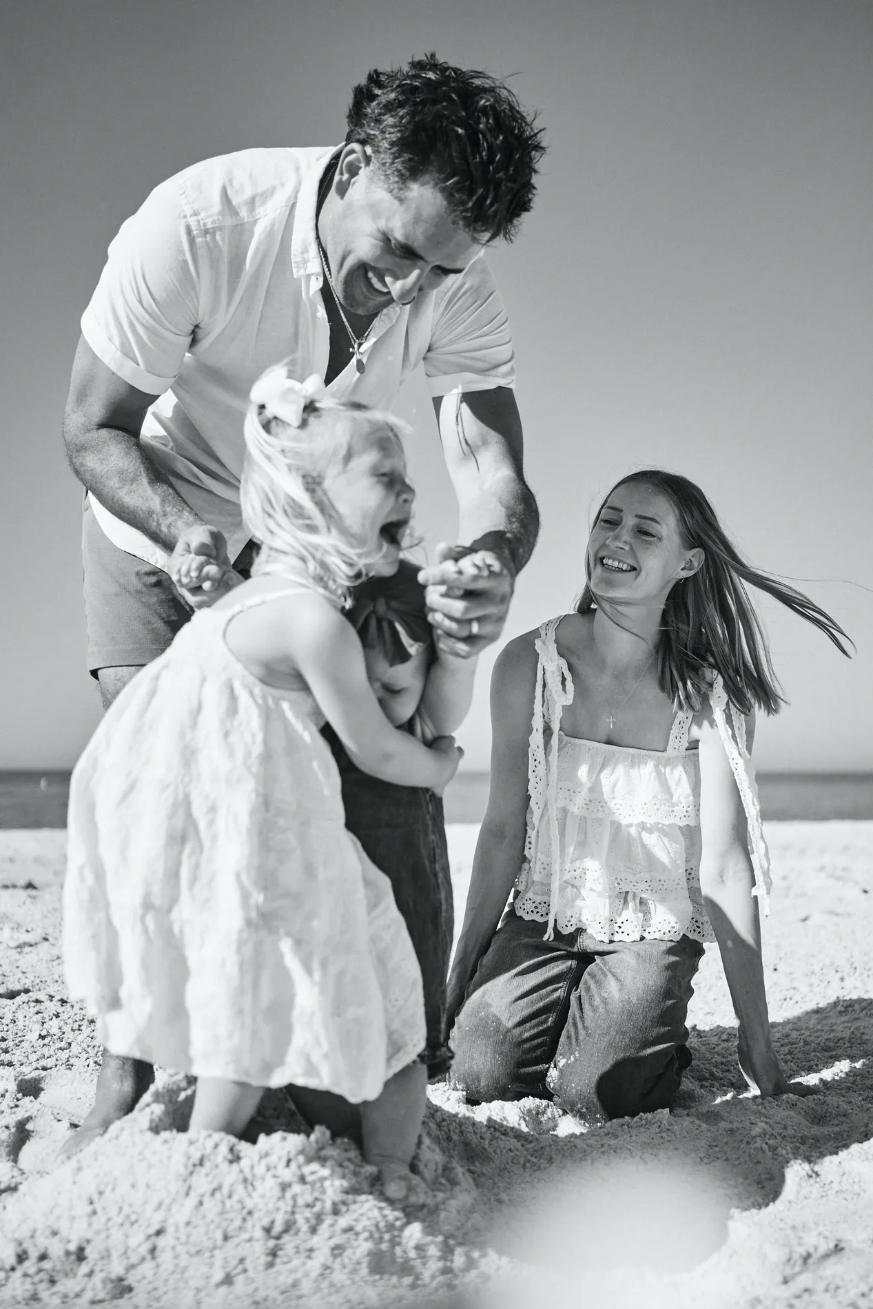 Black and white: a father lifting his laughing little daughter by the hands while the mother kneels on the sand and laughs, her hair blowing in the wind