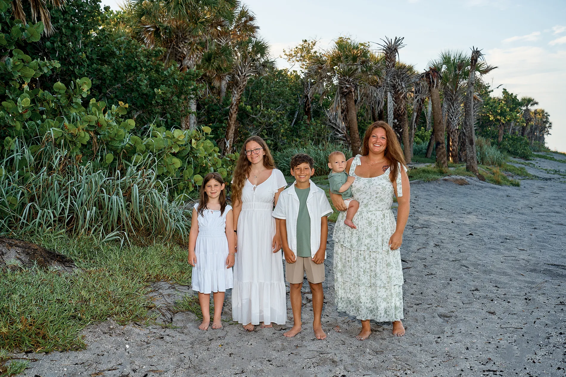 A woman holding a baby with a teenage girl, a boy and a young girl in white and sage green, standing barefoot on a sandy path under palms and sea grape at Caspersen Beach in Venice