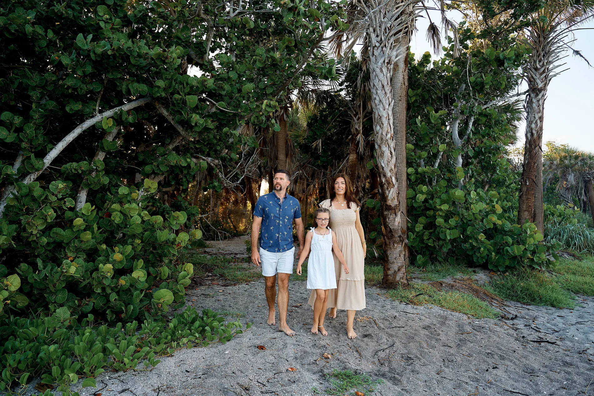 A father in a blue shirt, a mother in a long beige dress and their daughter in white standing hand in hand on a sandy path between palms and sea grape at Caspersen Beach in Venice