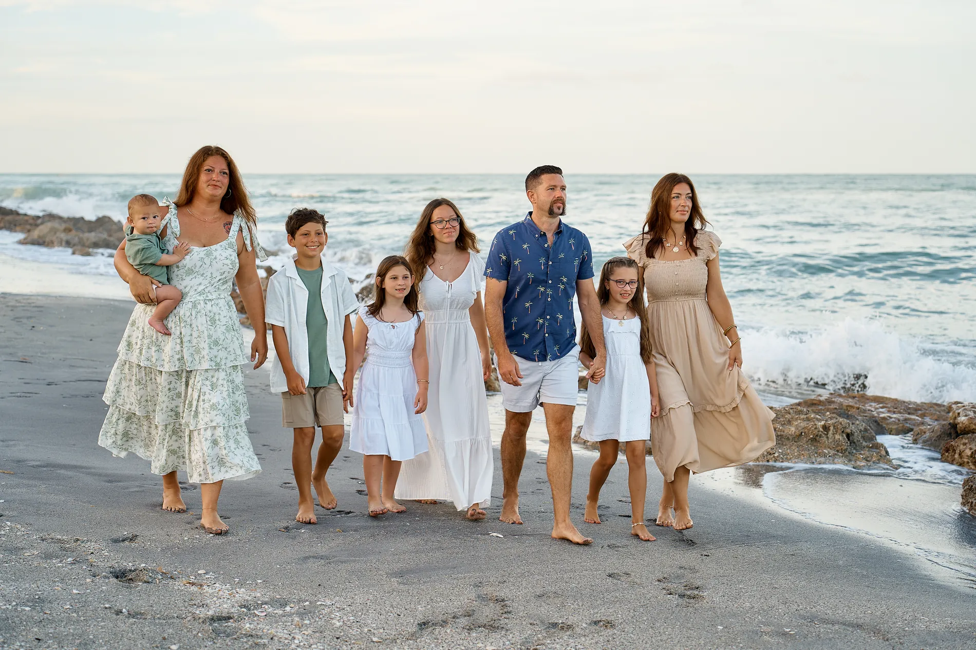 A family of seven and a baby walking along the dark sand at Caspersen Beach in Venice, rocks and a breaking wave behind them