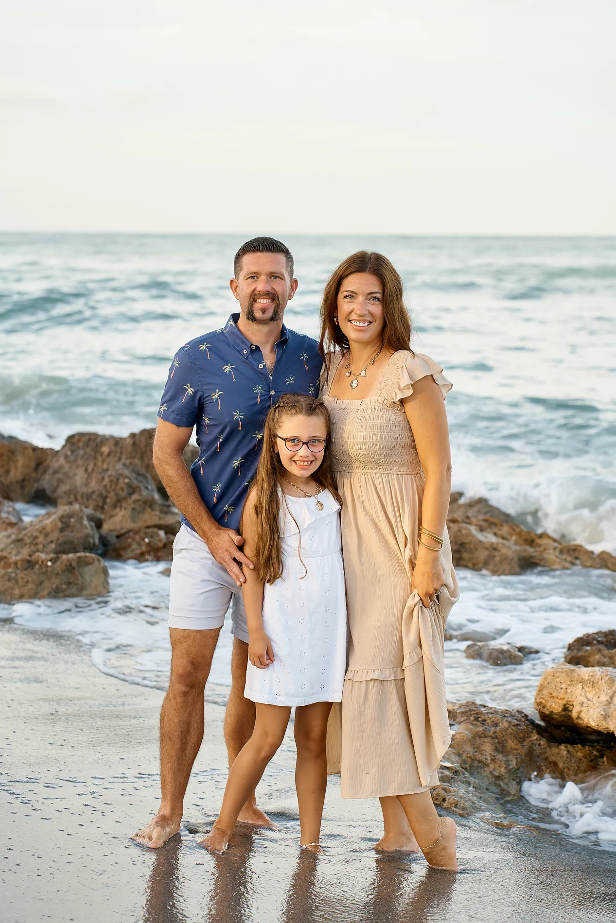 A father in a blue shirt, a mother in a beige dress and their daughter in white standing together at the waterline at Caspersen Beach in Venice, rocks and a breaking wave behind them