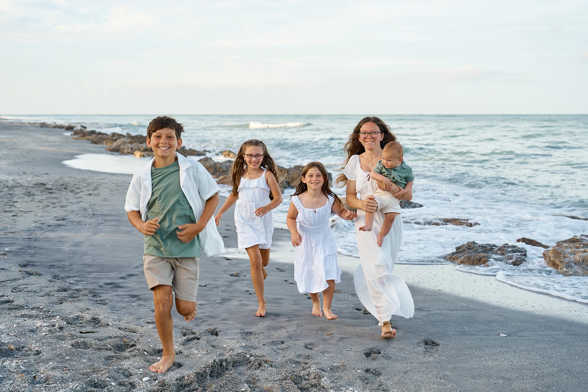 A mother in a long white dress carrying a baby, two girls in white sundresses and a boy in a sage shirt and khaki shorts running barefoot along the water at Caspersen Beach