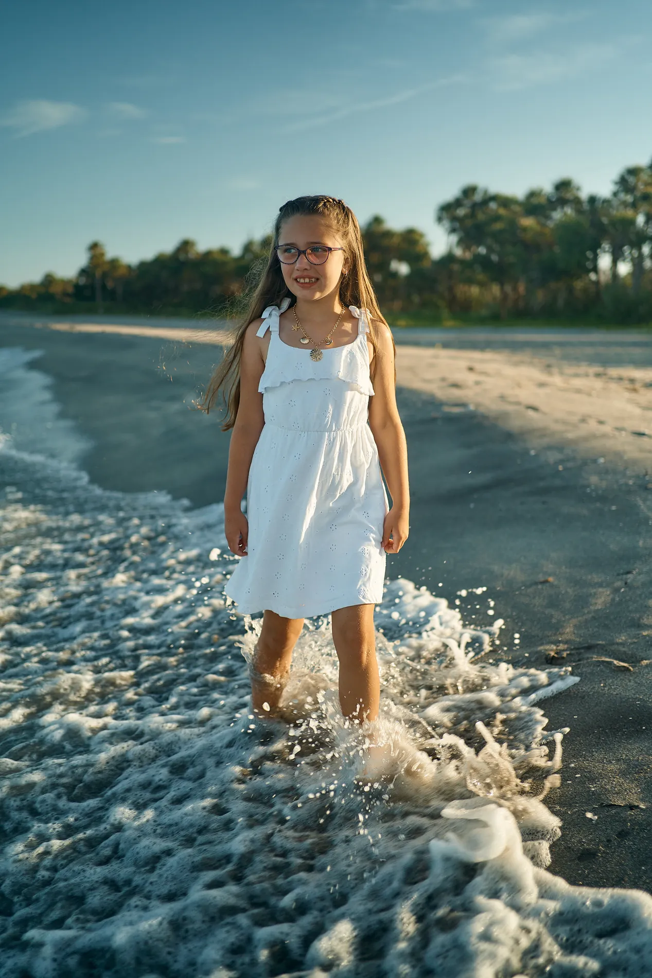 A girl in a white dress standing ankle-deep in the foam at Caspersen Beach, the curve of the shore and palms behind her