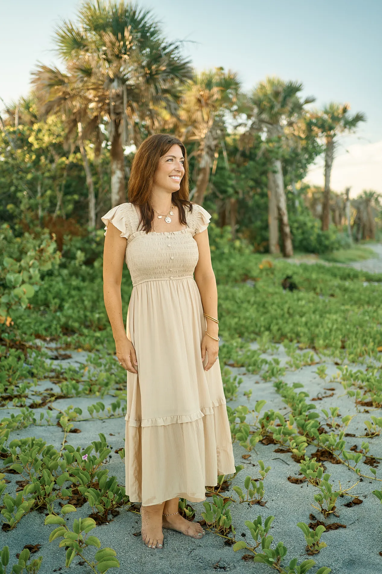 A smiling mother in a long beige dress standing barefoot on the sand among beach vines, palms and sea grape behind her at Caspersen Beach in Venice