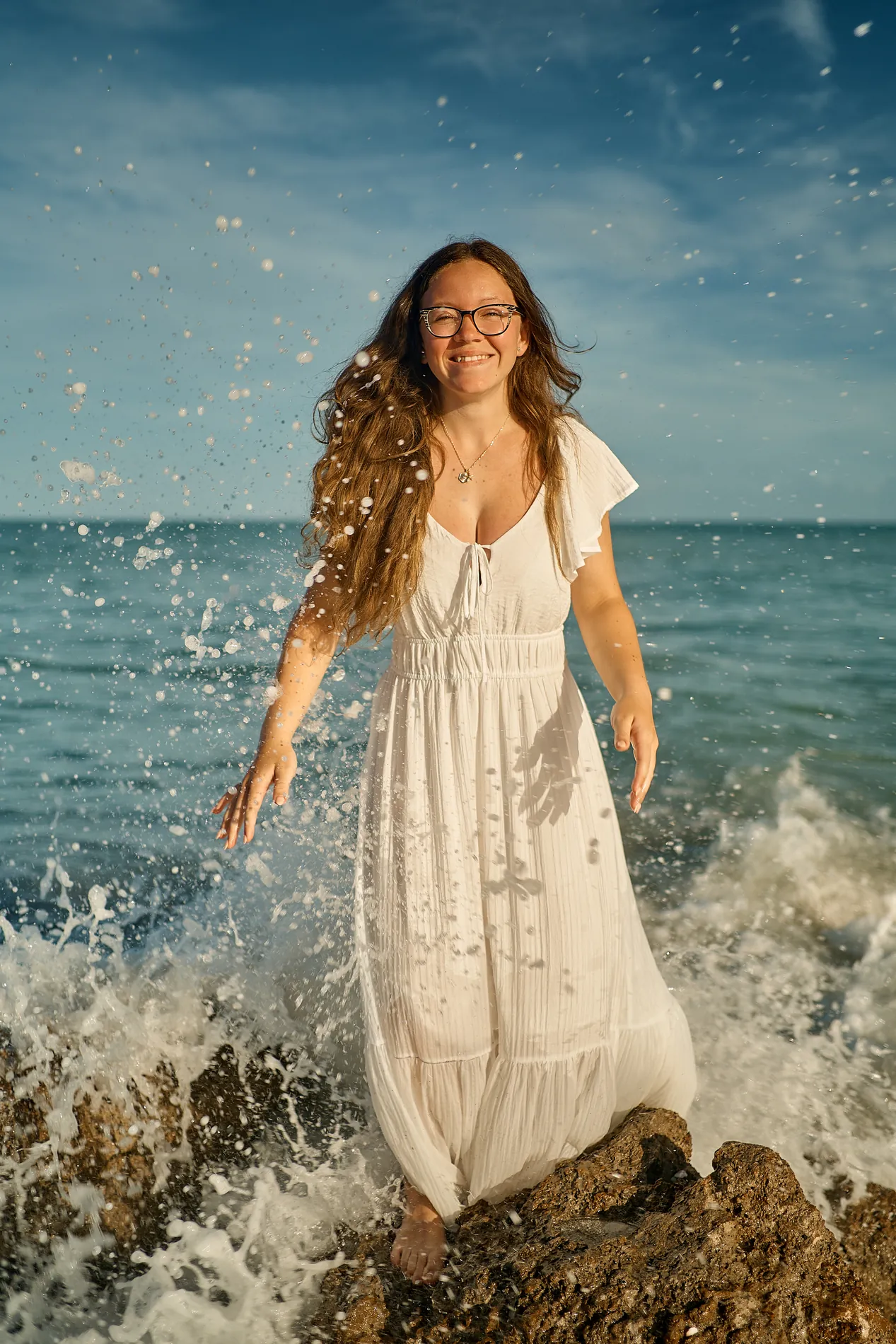 A woman in a white dress standing on a rock at Caspersen Beach in Venice as a wave breaks around her and throws spray into the air