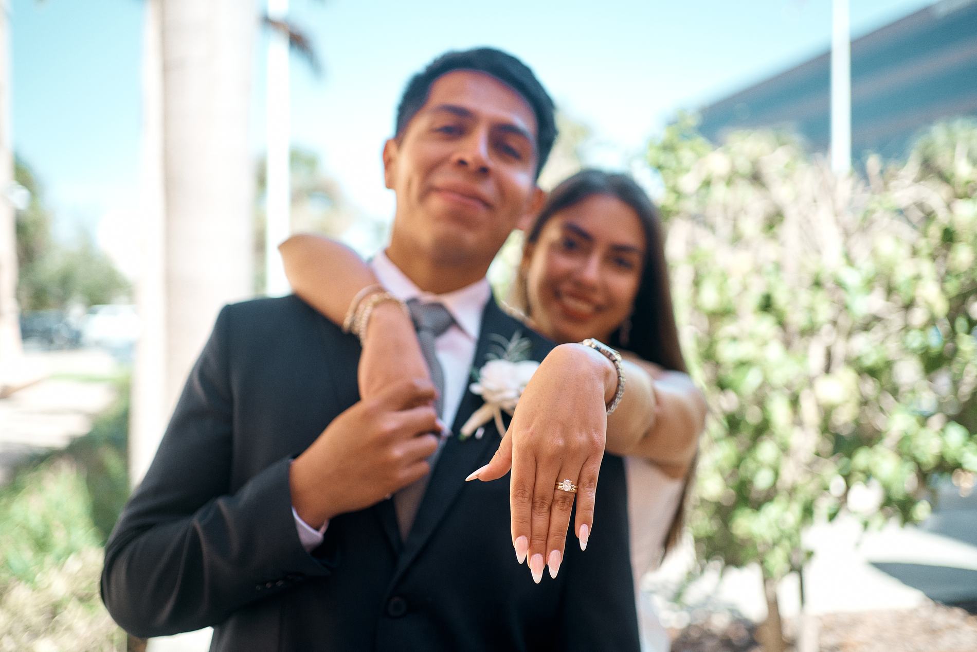A groom and bride smiling at the camera, her hand with the ring in front