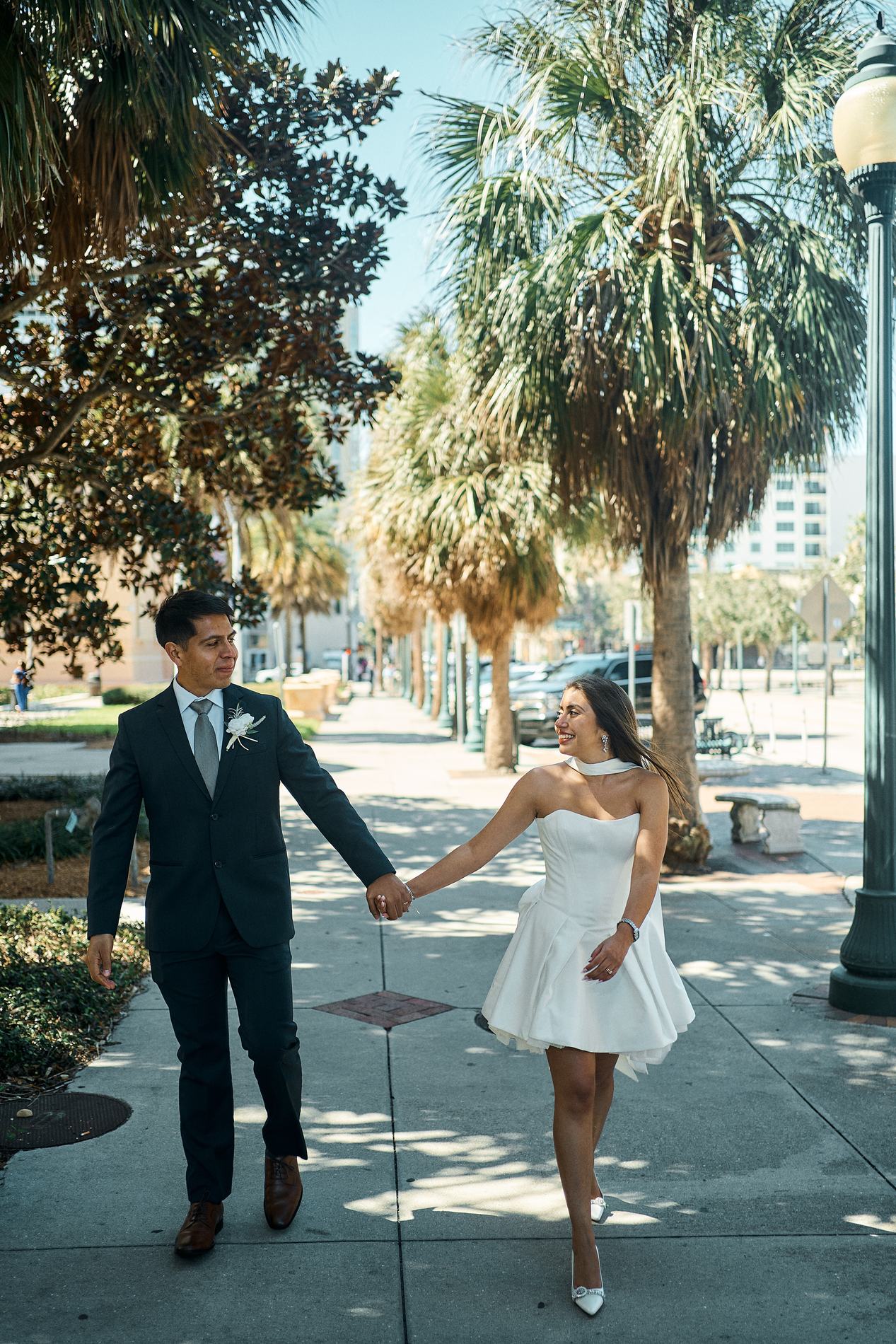 A bride and groom walking toward the camera on a downtown Sarasota sidewalk lined with palms