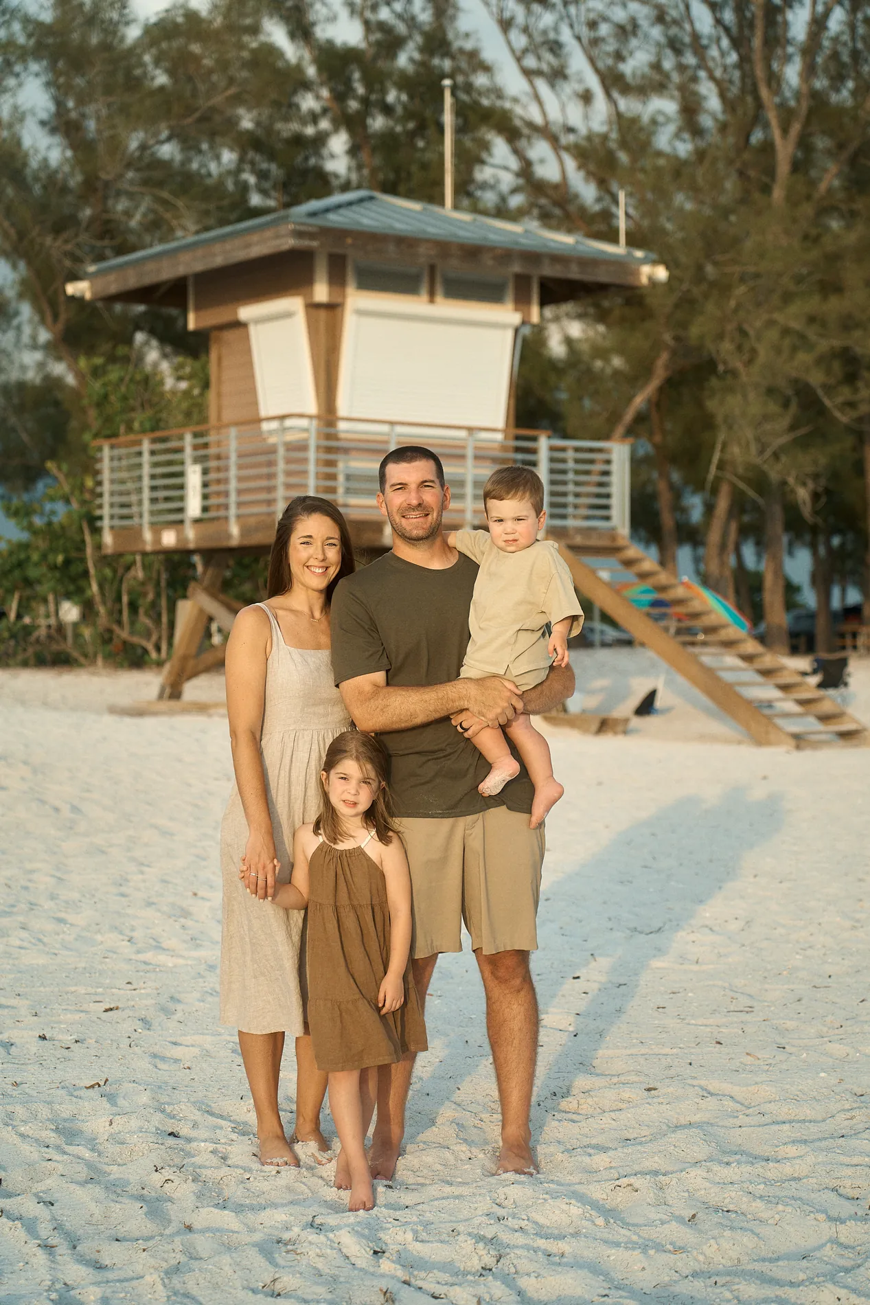 A mom and dad with their little girl and toddler son standing together on white sand in front of a wooden lifeguard tower at Coquina Beach, pines behind