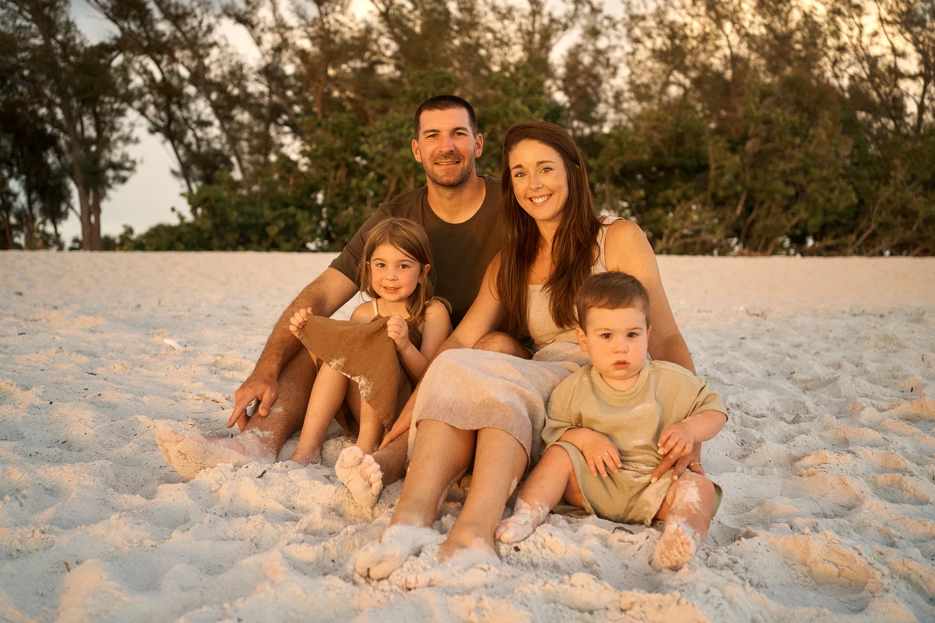 A mom and dad sitting on the white sand with their little girl and toddler son in golden evening light, pines behind them