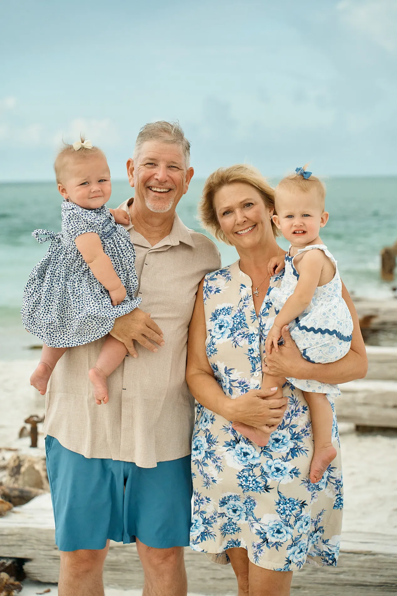 A laughing grandfather and grandmother on the beach, each holding a baby granddaughter in a blue dress, old wooden posts behind them
