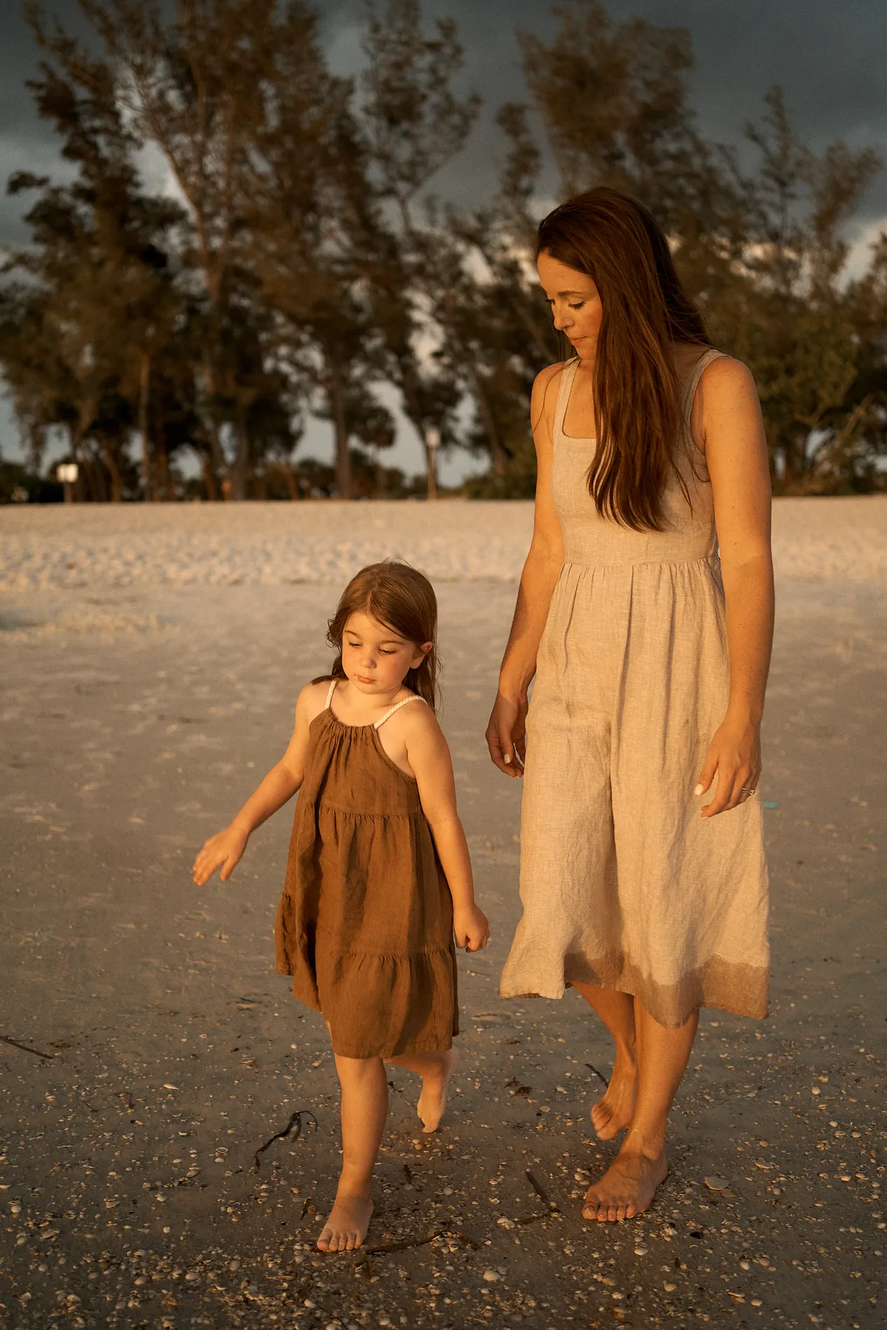 A mother and her little girl walking barefoot on the sand at Coquina Beach in low golden evening light, a dark storm sky and pines behind them