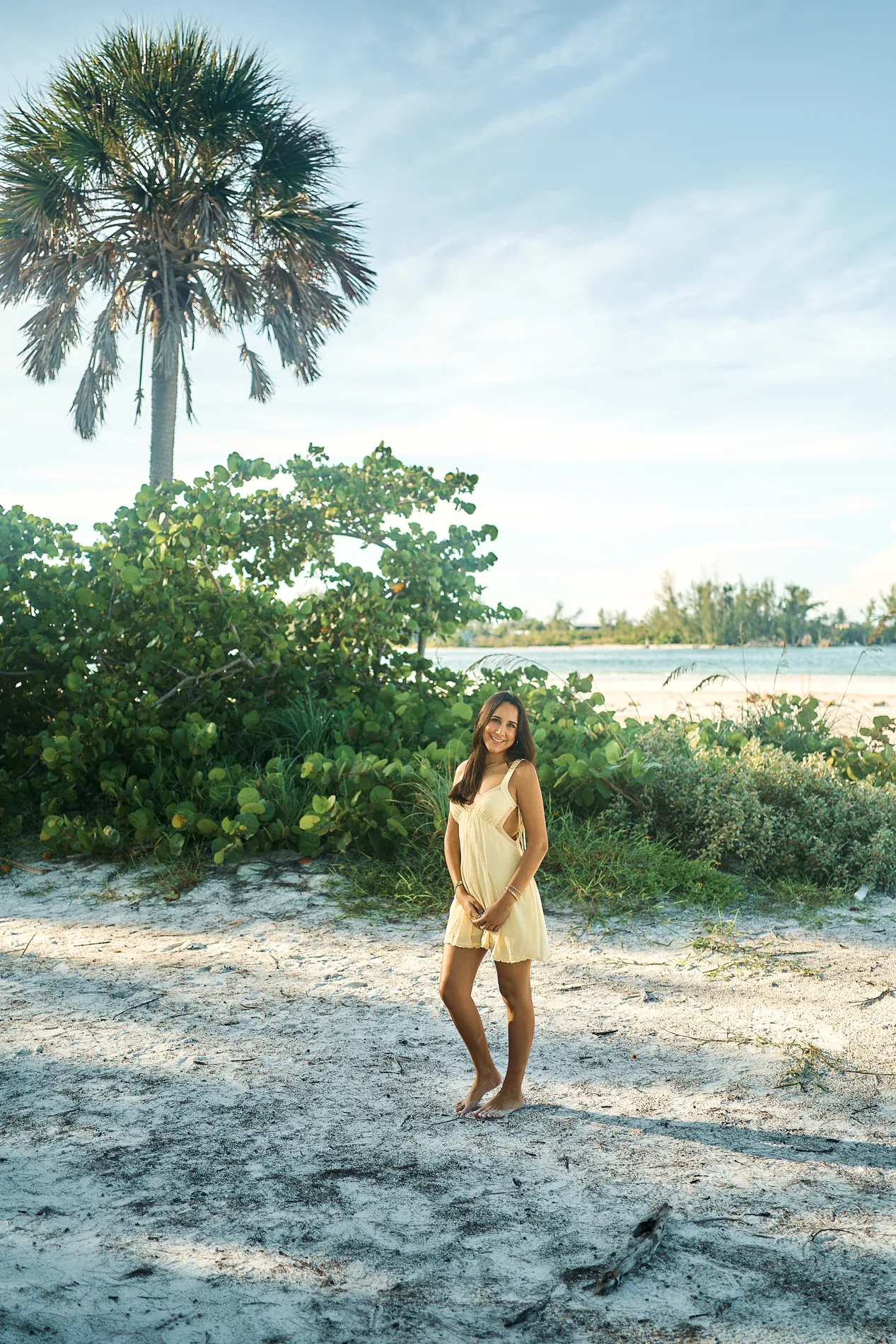 A young woman in a yellow dress standing barefoot on the sand in front of a cabbage palm and sea grape, the water behind her