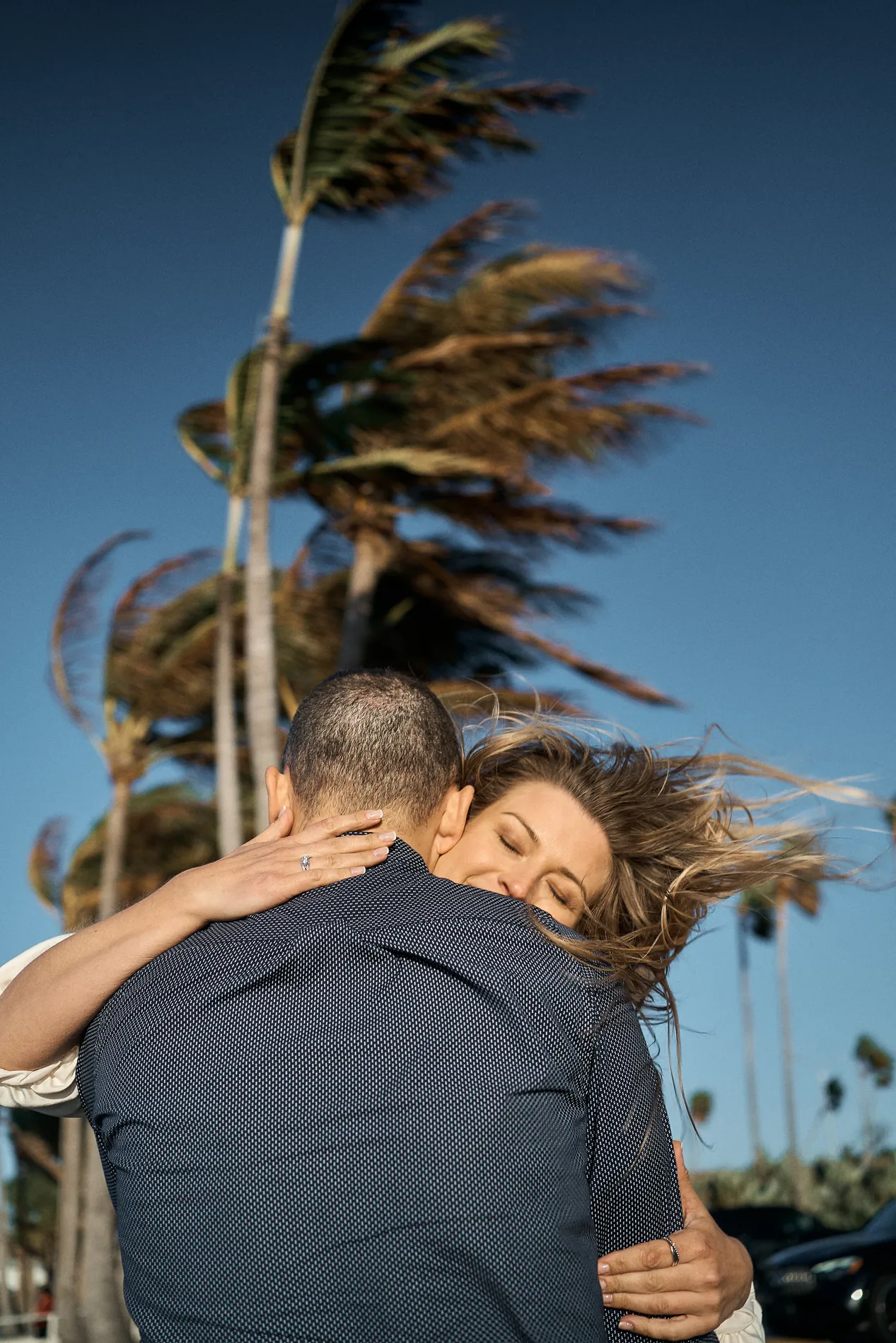 A couple holding each other tight on a very windy evening, her hair blowing across his shoulder, palm trees bending against a deep blue sky