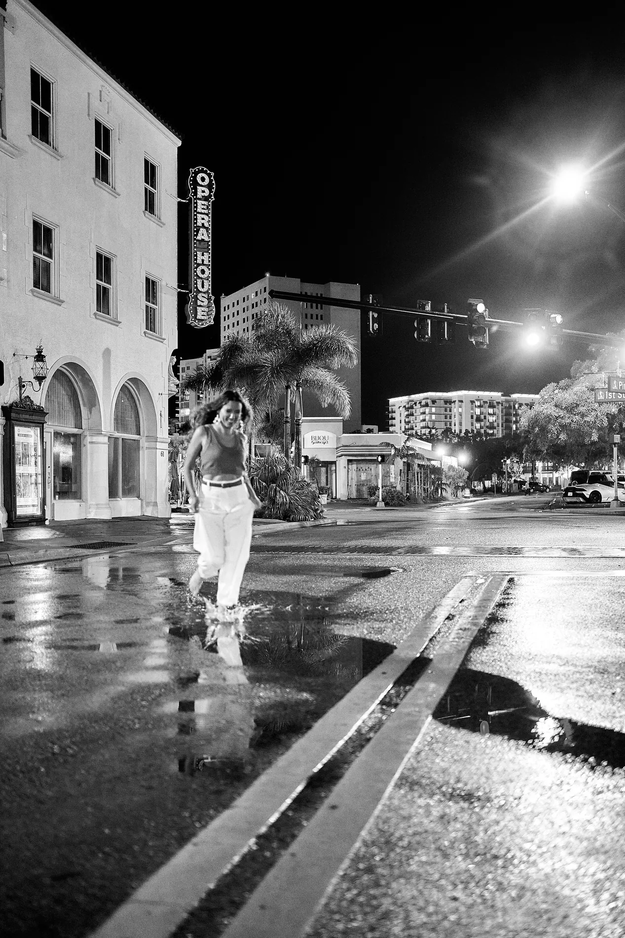 A young woman in white trousers running barefoot through a rain puddle at night in downtown Sarasota, the Opera House sign glowing behind her, black and white