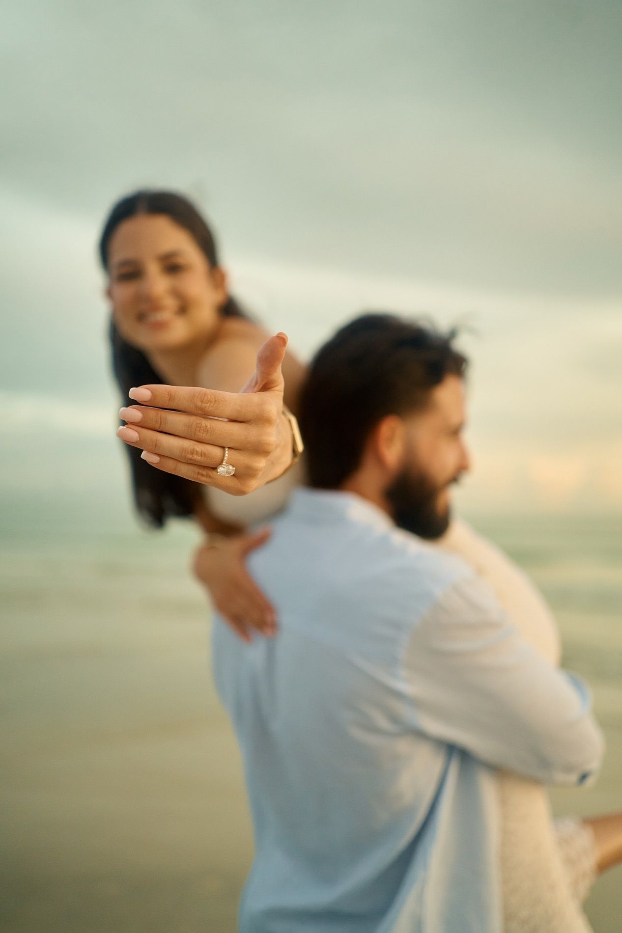 A couple after their proposal, the ring in focus, the beach behind them