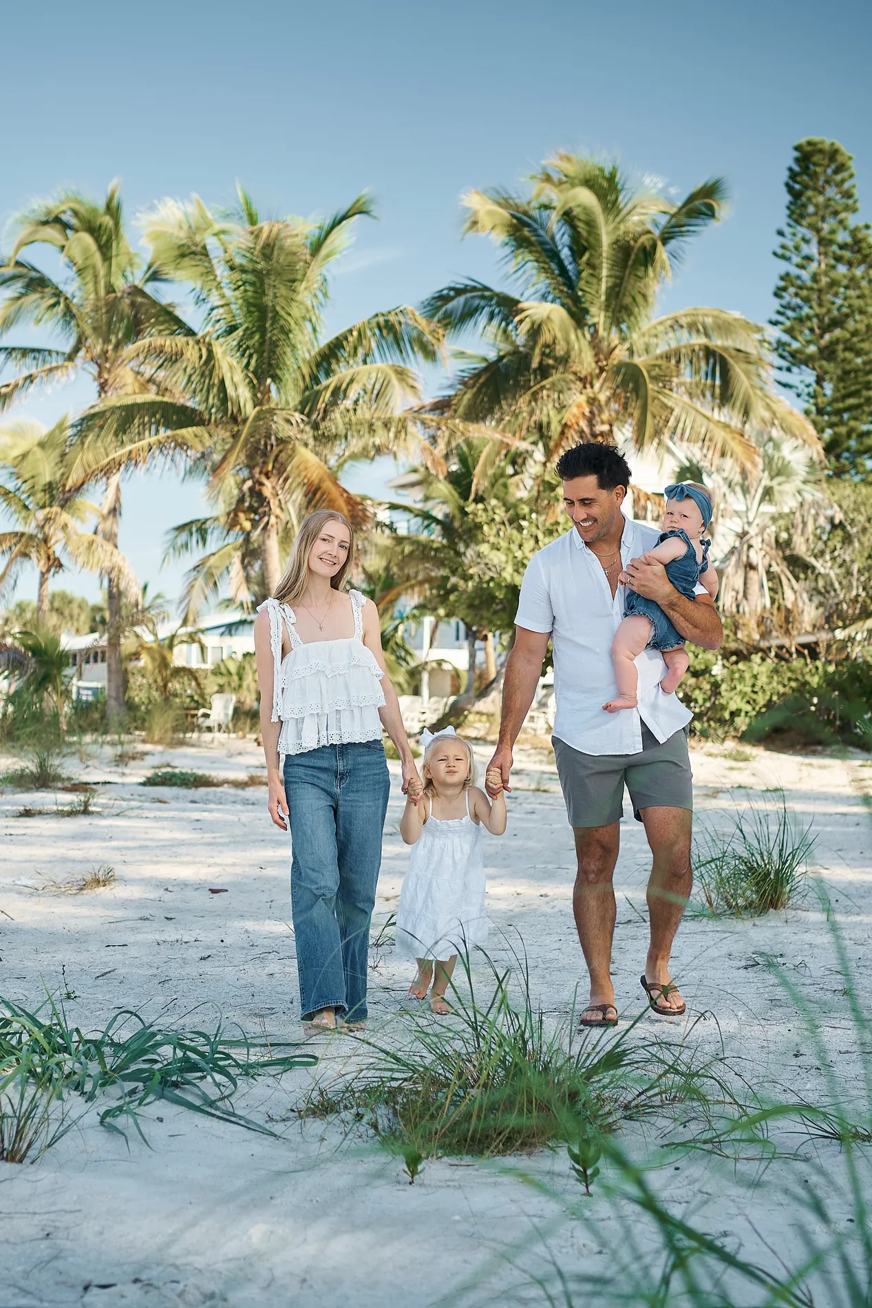A mother in a white top and jeans and a father carrying a baby walking a little girl in a white dress through dune grass, palm trees and a clear blue sky behind them