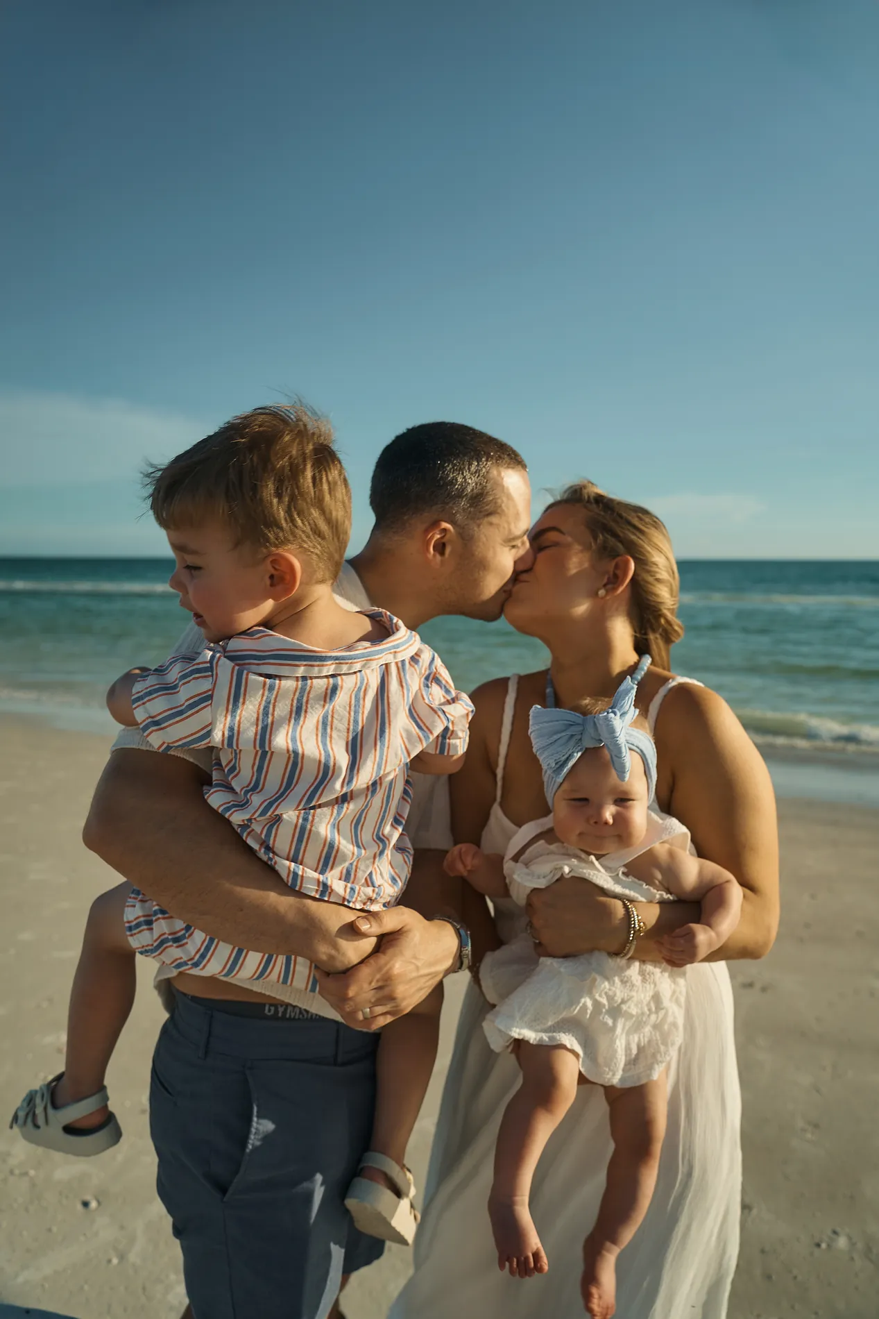 Parents kissing at the edge of the sea, the father holding a toddler in a striped shirt and the mother a baby with a blue bow