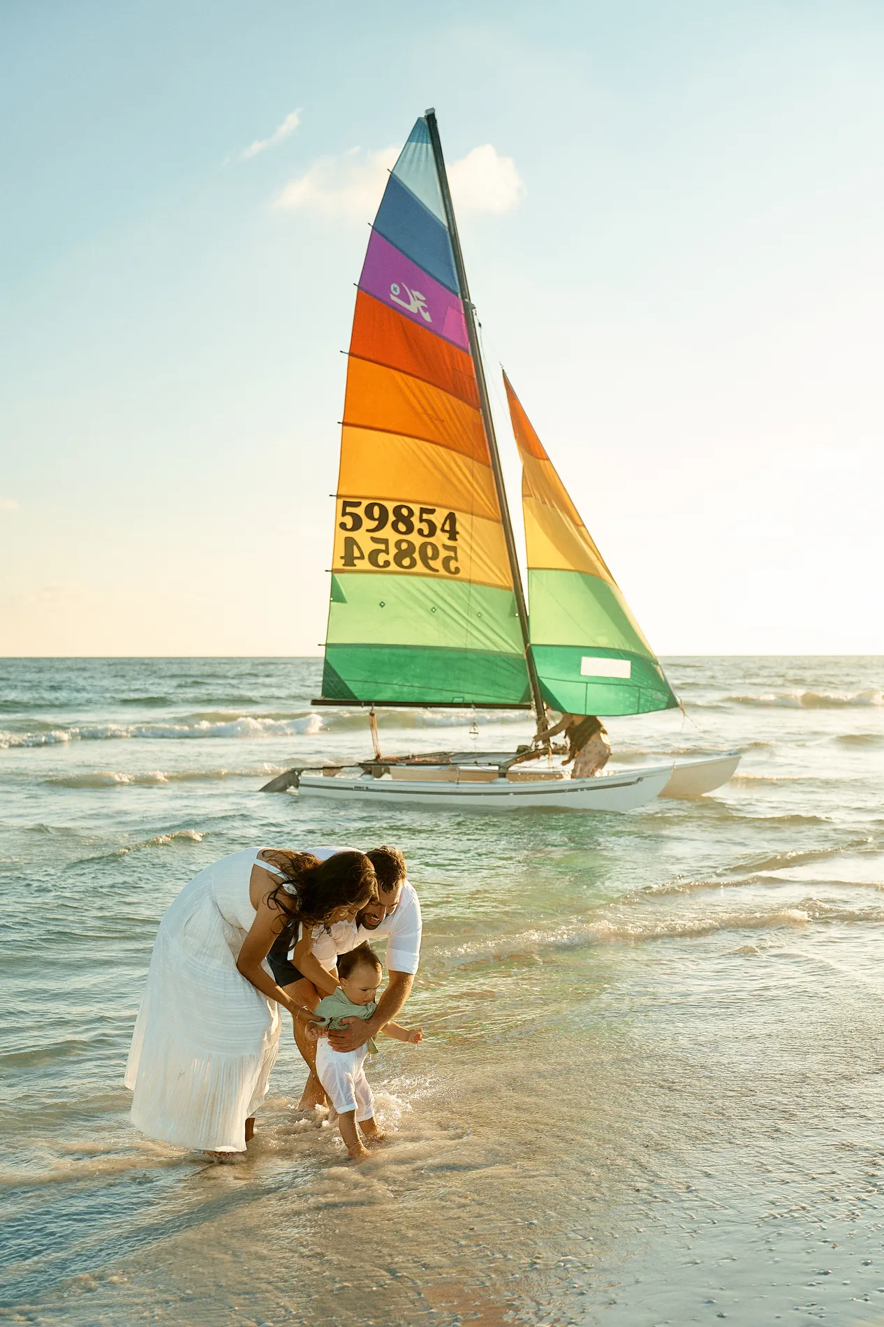 Parents holding up their baby to walk in the shallow water on Siesta Key under a pale, bright sky before sunset, a catamaran with a rainbow sail floating behind them with a sailor on board