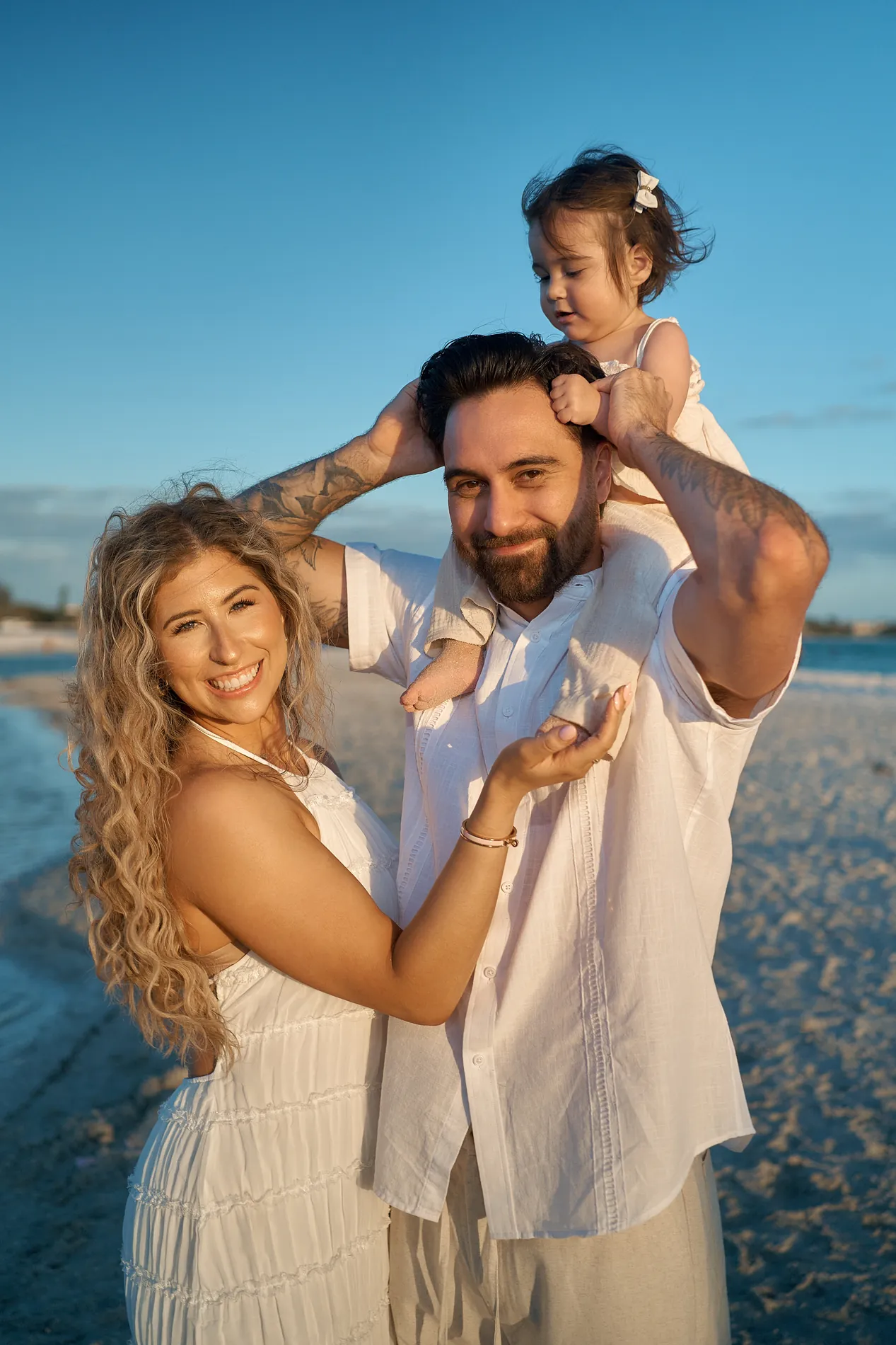 A mother laughing next to a father who holds his toddler daughter on his shoulders at the water line, lit by the low evening sun
