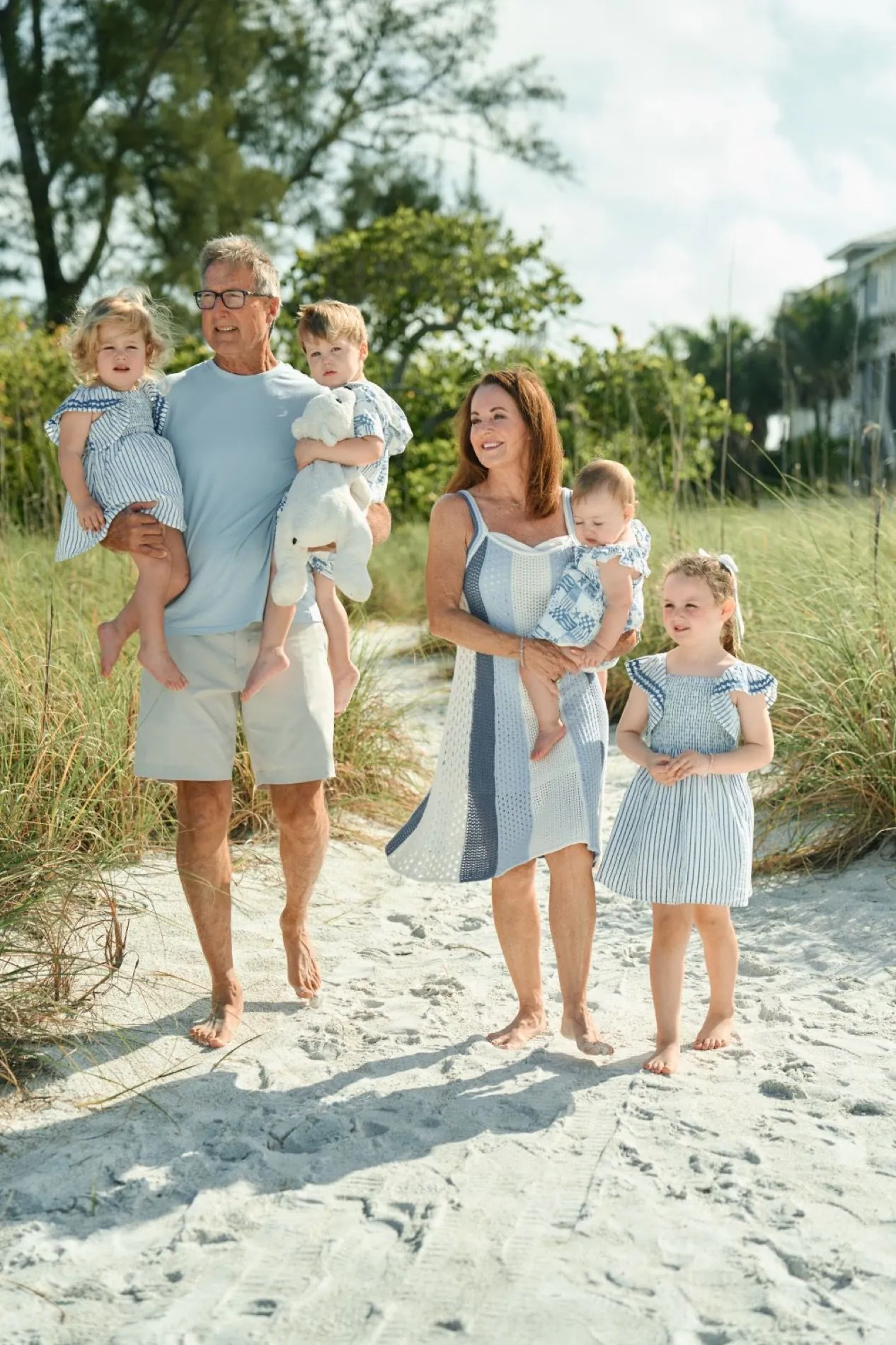 A grandfather in a light blue shirt carrying two little granddaughters on a sandy path through sea grass, their mother and a young girl beside him