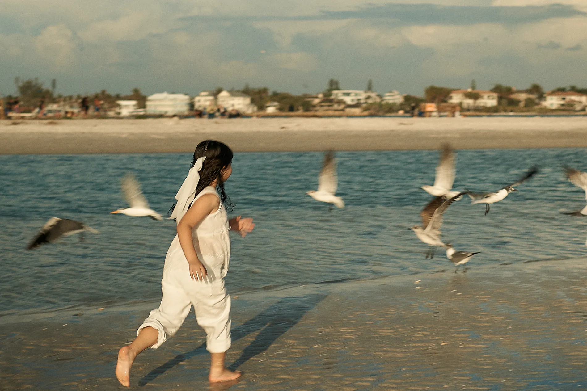 A little girl in a white romper, her long hair tied back with a white ribbon, running barefoot through the shallow water as seagulls take off around her