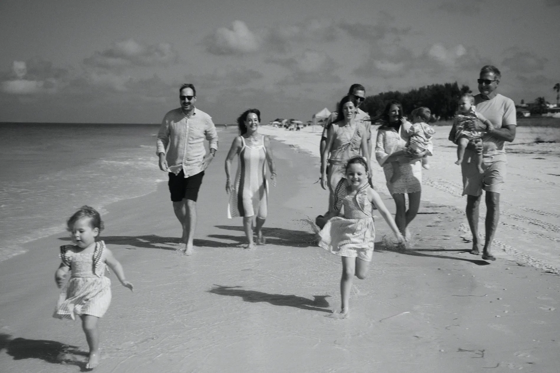 Black and white: a big family walking and running toward the camera on wet sand, a little girl in a dress out in front, an older girl laughing as she runs, a grey-haired man in sunglasses carrying a toddler