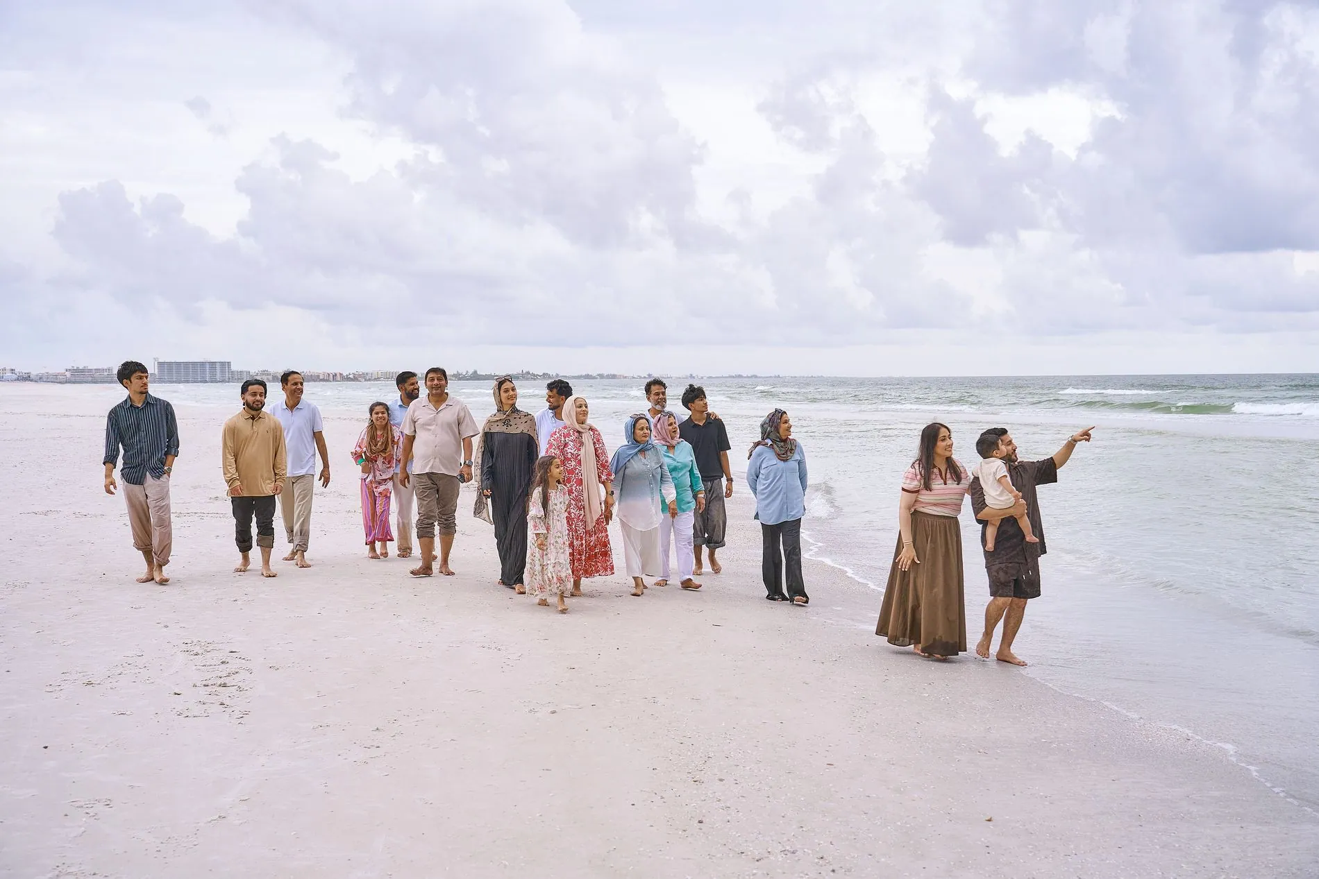 A big three-generation family, about eighteen people of all ages, walking together along the edge of the water on pale sand under a cloudy sky, a father at the front holding a toddler and pointing ahead