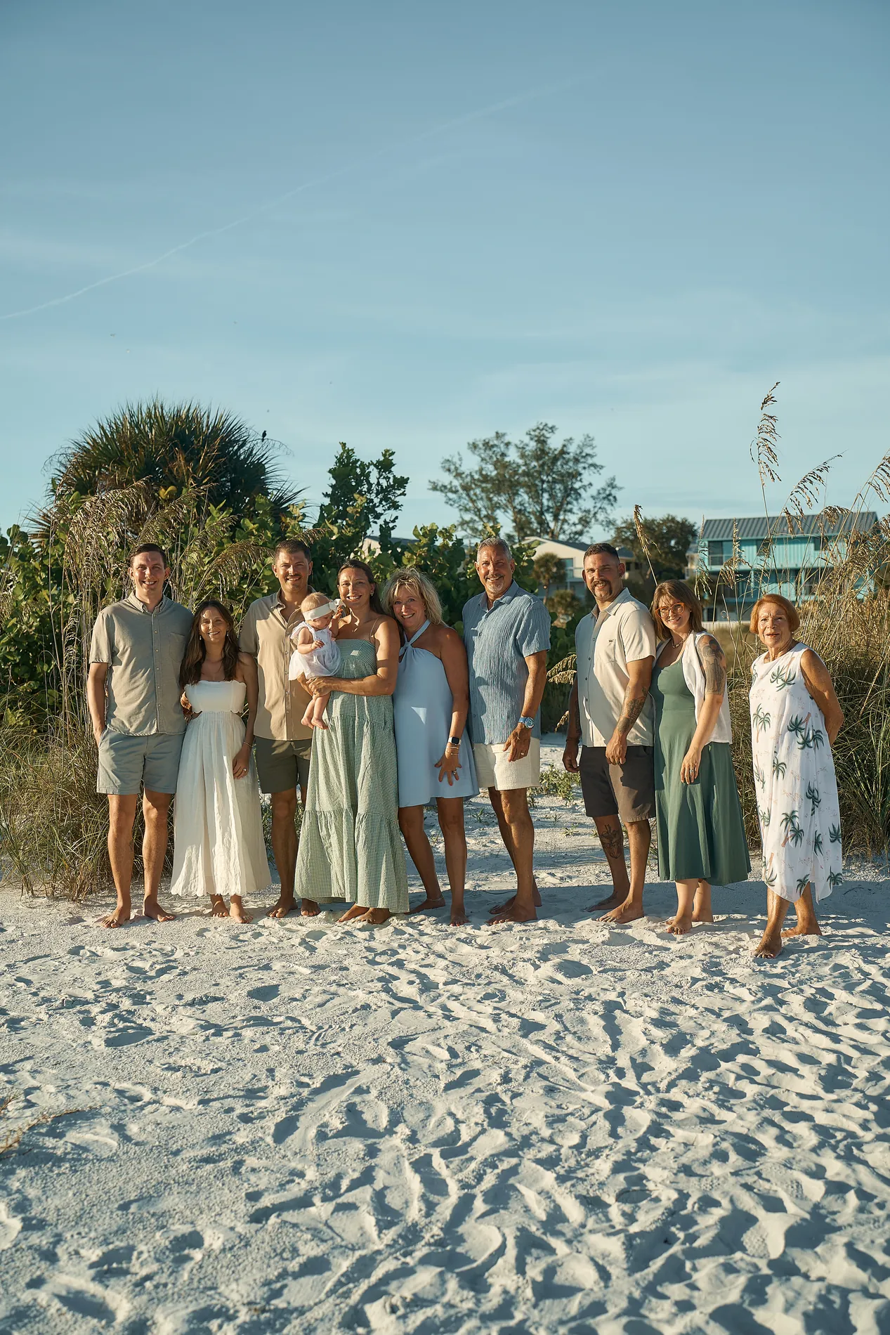 Ten adults and a baby standing in front of sea grapes and sea oats: sage, olive and pale blue dresses, a cream dress, men in grey, blue and white shirts, a grandmother in a white dress with a green palm print, a baby in the middle