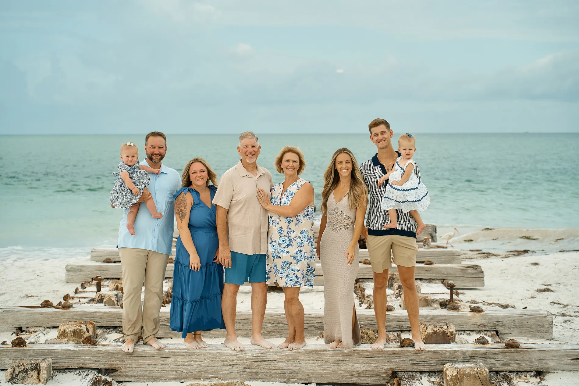Six adults and two baby girls standing on the old wooden pilings of a pier by turquoise water: a woman in a blue dress, men in a light blue shirt with khaki trousers, a beige shirt with blue shorts and a striped knit polo, a woman in a blue floral dress, another in a long beige knit dress, the babies in white and blue carried by two of the men